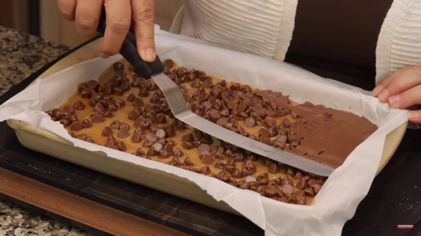 In this indoor image, a person is using an offset spatula to spread melted chocolate chips over a layer of baked goods in a rectangular baking dish lined with parchment paper. The dish is resting on a wooden surface, likely a kitchen countertop. The chocolate chips are partially melted, suggesting they have been heated, and the person is smoothing them into an even layer across a light brown base, possibly a type of cookie or bar. The hands are visible, with one hand holding the edge of the parchment paper and the other gripping the spatula firmly. The lighting is warm and inviting, accentuating the glossy sheen of the melted chocolate. The scene conveys a sense of hands-on baking, with an emphasis on the process of preparing a sweet treat. There are no visible text or branding elements in the image.