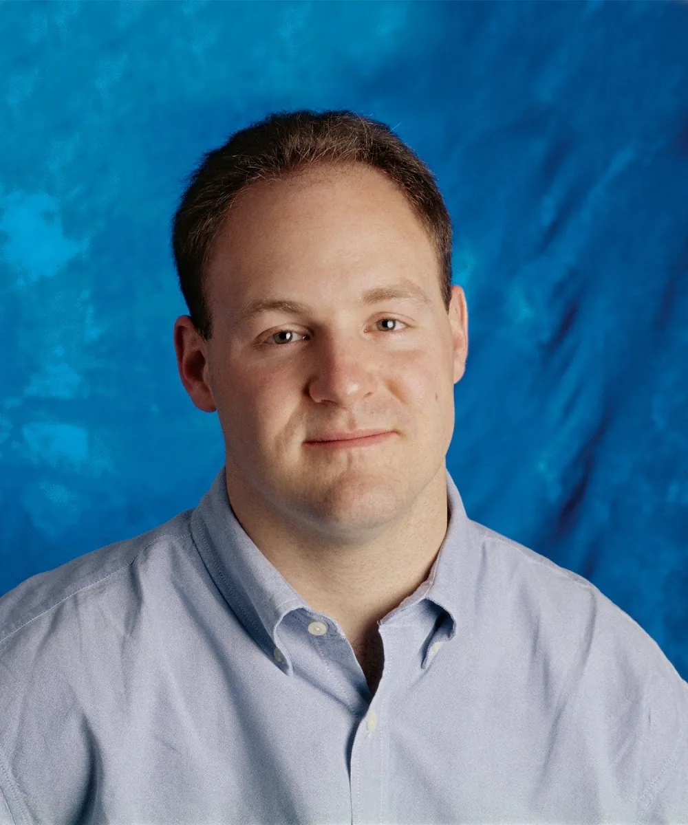 The image is a professional portrait of Daniel Lewin. He is situated against a vivid blue backdrop that provides a vibrant contrast to his light skin tone. Daniel is wearing a pale blue button-up shirt, which complements the background color, creating a harmonious color scheme. The shirt is neatly buttoned up to the collar, giving a formal and polished appearance. He has short, neatly groomed hair and a slight smile, conveying a calm and approachable demeanor. The lighting is soft and even, highlighting his facial features without harsh shadows. The photograph is likely taken in a studio setting, indicated by the uniform background and controlled lighting, suggesting a formal or professional purpose for the image.
