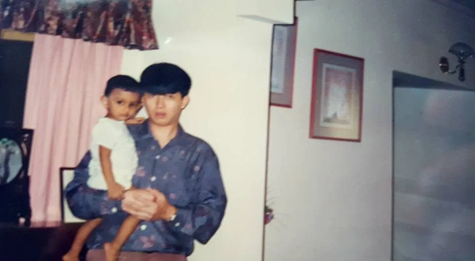 The image features two individuals indoors. On the left, a young child, likely Lehan Edirisinghe, is being held in the arms of an adult man, suggested to be his "fake Asian dad," Martinn Teng. The man is wearing a patterned dark shirt and has his hair styled in a classic, neat fashion. The child is dressed in a simple light-colored outfit, and both individuals are looking towards the camera with neutral expressions.
The setting appears to be a living room, as indicated by the visible decor. There is a pink curtain and a wooden frame part of a door or window in the background. Additionally, framed artwork is hanging on the wall, adding to the homely ambiance. The lighting is soft, suggesting a casual, warm atmosphere typical of family gatherings. The photograph has the appearance of a personal, candid snapshot, capturing a moment of family connection.