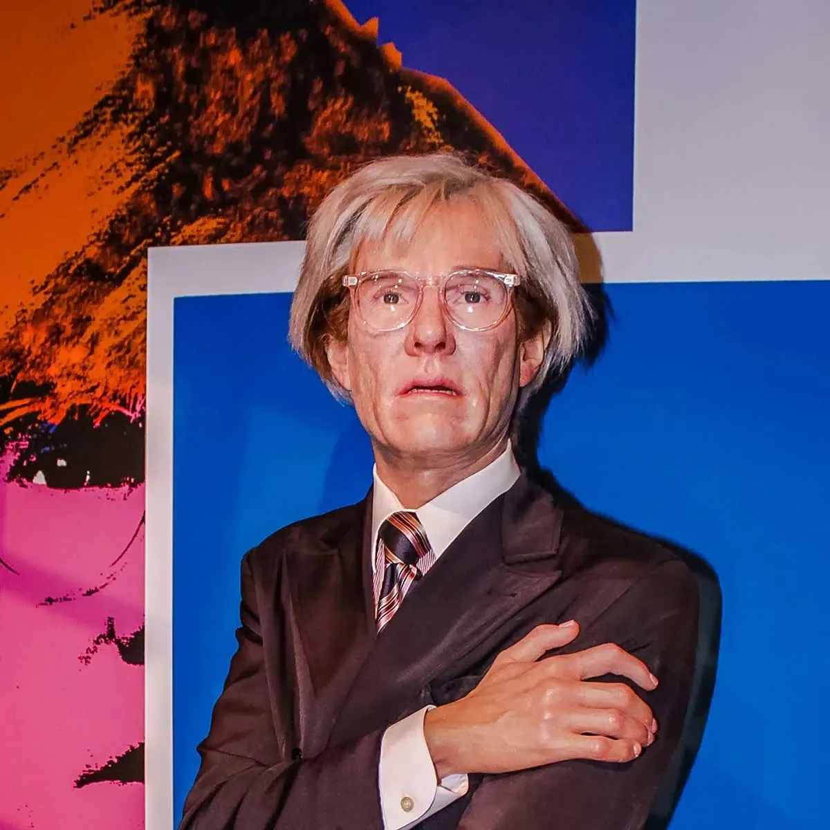 ```
The image features a lifelike representation of Andy Warhol, a prominent figure in the pop art movement. The setting appears to be an indoor exhibition, possibly a museum or gallery, given the vibrant backdrop and artistic context. Warhol is depicted with his characteristic silver hair and is wearing clear-framed glasses. His attire consists of a dark suit, white shirt, and a striped tie, aligning with the formal yet artistic persona he was known for.
The background is colorful, featuring bold hues of blue, orange, and pink, reminiscent of Warhol's iconic art style. The composition suggests a focus on Warhol's influence on modern art, highlighted by the vivid colors and striking visual elements around him. The expression on Warhol's face is neutral, with a slight hint of introspection, capturing his enigmatic and contemplative nature. The lighting is bright, emphasizing the subject and adding to the overall vividness of the scene.
