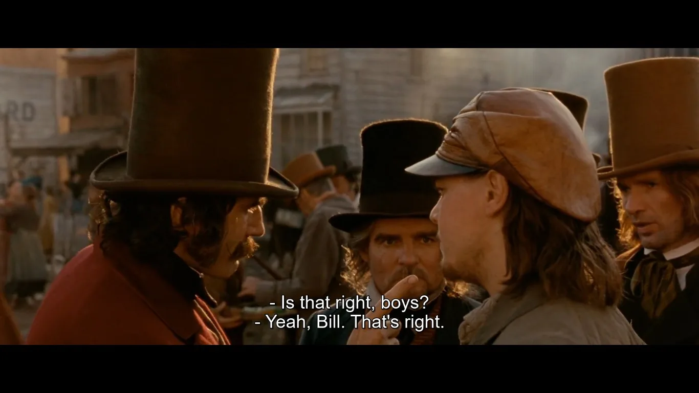 In this scene from "Gangs of New York," a group of men is engaged in a conversation on a bustling 19th-century street. The man on the left, identified by the subtitles as Bill, wears a tall black top hat and a dark coat, exuding a commanding presence as he addresses the others. Surrounding him are several men in period attire, including top hats and weathered coats, characteristic of the film's historical setting. The man in the center, facing Bill, affirms his statement with a serious expression. The lighting is warm, casting an authentic historical glow that complements the earthy tones of the costumes and the wooden structures in the background. The street is lively, with blurred figures indicating movement and activity, enhancing the scene's immersive period atmosphere. The subtitles present a snippet of the dialogue, emphasizing the authority and respect commanded by Bill.