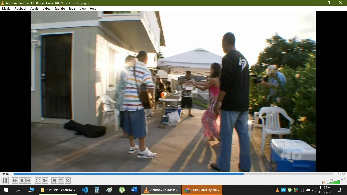 The image is a screenshot from the VLC media player, displaying a scene from "Anthony Bourdain No Reservations S04E09." The setting is outdoor with a casual, festive atmosphere. A group of people is gathered near a house with a canopy tent in the background. The lighting suggests it is late afternoon or early evening, with sunlight casting long shadows on the ground. Individuals are casually dressed; one person is playing a guitar, contributing to the relaxed mood. Plastic chairs and a cooler are visible, indicating a laid-back gathering or party. The focus seems to be on the interaction and cultural exchange, fitting the theme of Anthony Bourdain's travel and cooking show. The screenshot includes the VLC media player interface with controls and options visible at the top and bottom of the screen.