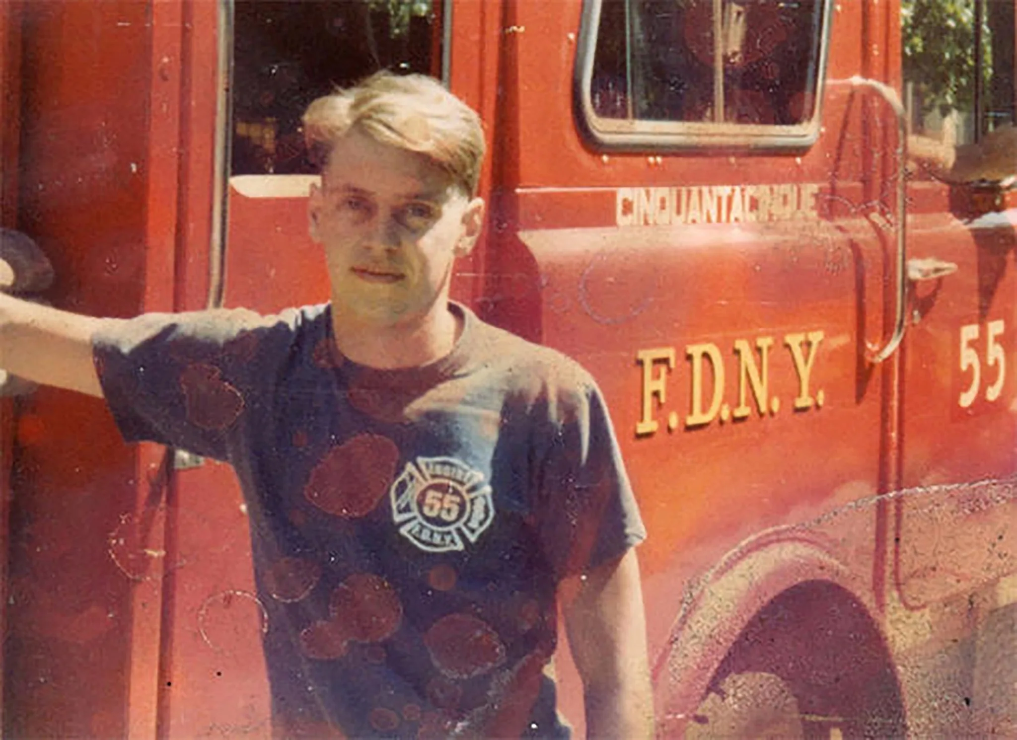 The image features Steve Buscemi standing next to a red FDNY fire truck. He's wearing a navy blue T-shirt with a firefighter emblem featuring the number 55, indicating his affiliation with the fire department. The fire truck behind him also displays "F.D.N.Y. 55" in bold yellow letters. Steve Buscemi is casually leaning against the truck with his left arm, giving off a relaxed and approachable demeanor. His expression is neutral, and his hair is swept to the side. The overall lighting suggests it is daytime, casting natural light across the scene. The truck's vibrant red contrasts with Buscemi's darker attire, drawing attention to both the man and the vehicle. This image captures a moment from Buscemi's past, emphasizing his connection to the fire department.