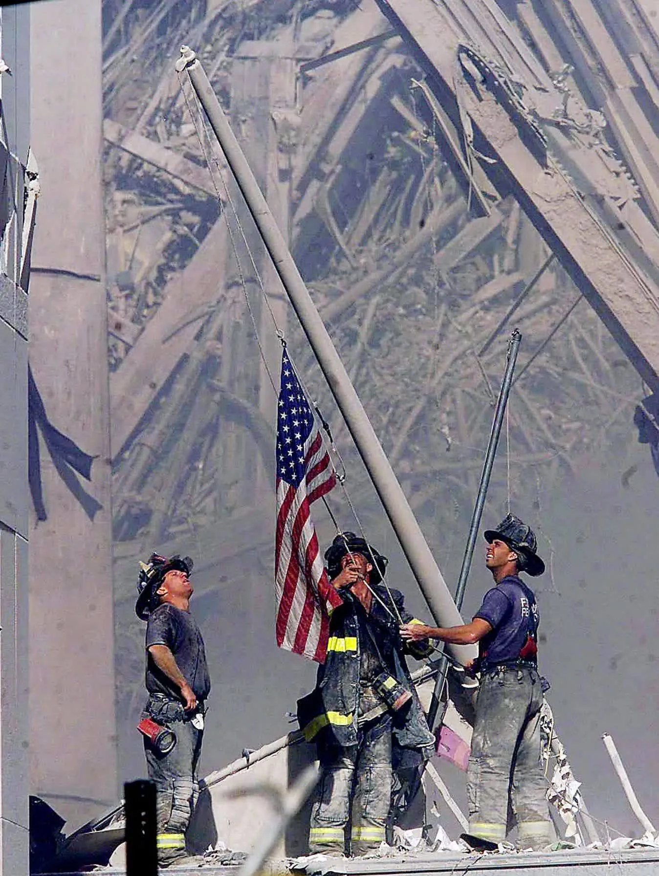 This image depicts three firefighters raising an American flag amidst the rubble and debris of a destroyed building. The scene is heavily covered in dust, with broken beams and twisted metal in the background, indicating the aftermath of a disaster. The firefighters stand resolute, wearing helmets and uniforms coated with dust. One firefighter is holding the flagpole steady while another adjusts the flag, and the third looks on, their expressions somber yet determined. The American flag is a focal point, symbolizing resilience and unity. The lighting is natural, casting shadows and highlighting the devastation surrounding them. This moment is captured with a sense of hope and defiance in the face of tragedy, emphasizing the bravery and dedication of first responders.