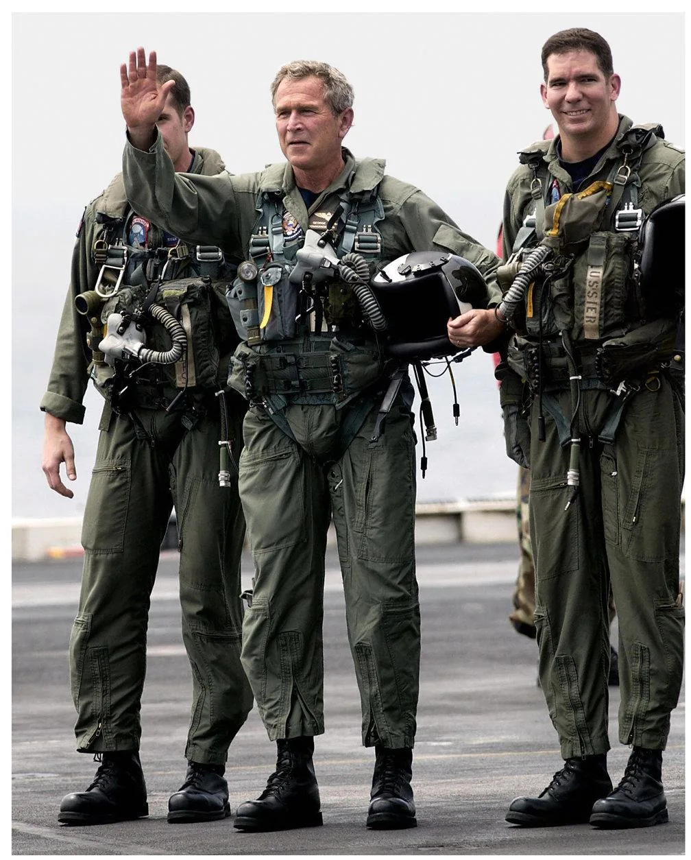 ```
George W. Bush is depicted in this image standing on what appears to be the deck of an aircraft carrier. He is flanked by two other individuals, all dressed in military flight suits. George W. Bush, positioned in the center, is raising his right hand in a waving gesture. His expression is serious but approachable, fitting the setting of a public appearance or official visit. The flight suit is olive green, equipped with various straps and gear typical for aviation, including a helmet held in his left hand. The individuals beside him are similarly attired, suggesting a shared purpose or event, likely related to military or governmental operations. The background is slightly blurred, indicating the vastness of the carrier deck extending towards the horizon under an overcast sky. The mood is formal yet friendly, with the setting emphasizing military readiness and leadership.