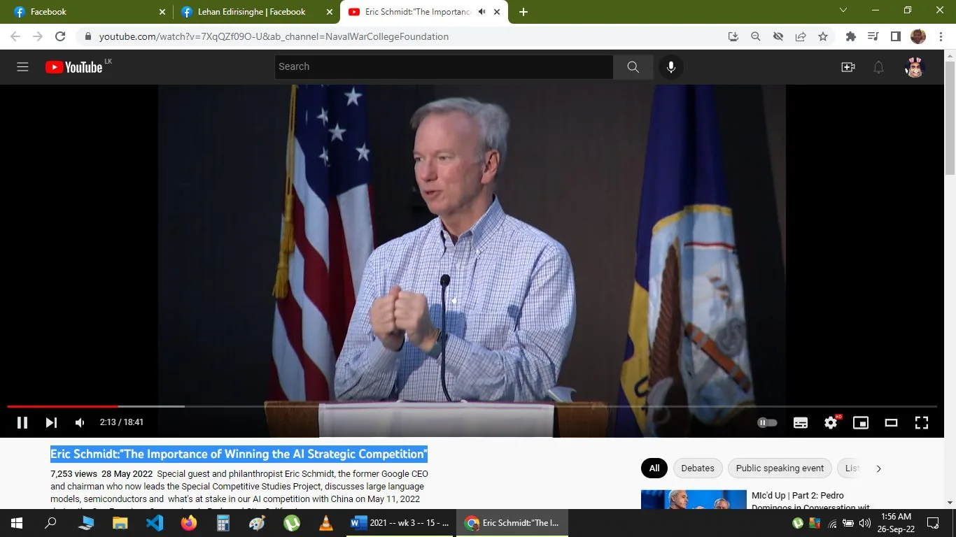 The image is a video frame from a YouTube presentation titled "Eric Schmidt: 'The Importance of Winning the AI Strategic Competition'" on the NavalWarCollegeFoundation channel. Eric Schmidt, identified in the text as the former Google CEO, is speaking at a podium. He is wearing a checkered shirt and appears engaged and expressive as he delivers his talk. The setting is indoors, with American and possibly naval flags visible behind him, indicating a formal or governmental venue. The lighting is soft, focusing on Schmidt, who is centered in the frame. The video description mentions the event took place on May 11, 2022, where Schmidt discussed topics like large language models, semiconductors, and AI competition with China. There are no captions or subtitles visible on this frame, but the context suggests a focus on strategic technological advancements and competitive studies.