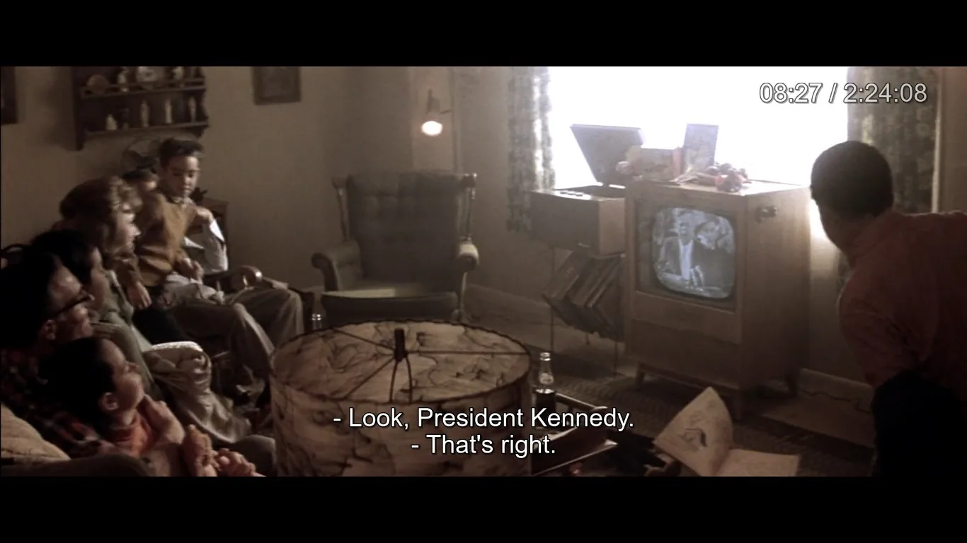 The image depicts a cozy, vintage living room scene where a family is gathered around an old-fashioned television set. The room is warmly lit, with soft, natural light coming through a window with floral curtains. In the center of the room, there's a round table with a decorative lampshade featuring a marbled design. The family, including adults and children, is seated on a couch and armchairs, intently watching the television. The TV screen displays a black-and-white image, and the caption reveals they are watching "President Kennedy." This suggests a historical or retro setting, possibly from the early 1960s. The room is furnished with a mix of upholstered chairs and wooden furniture, and a shelf on the wall holds various knick-knacks, adding to the lived-in feel of the space. The atmosphere conveys a sense of togetherness and engagement with a significant broadcast event.
