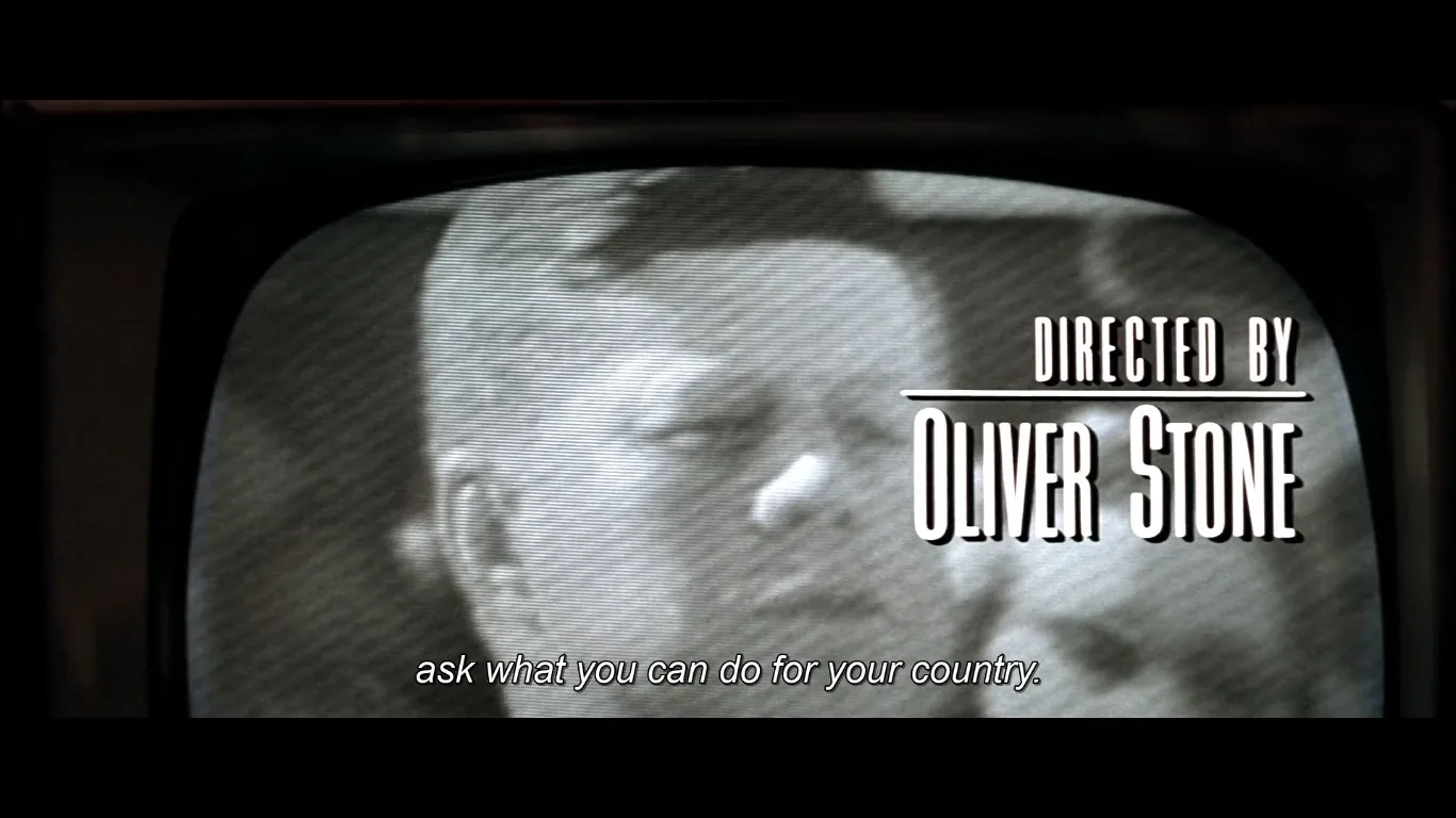 This image is a black and white video frame from the film "JFK," directed by Oliver Stone. The frame shows a close-up of a television screen displaying a historical broadcast. The screen is filled with horizontal lines typical of older television footage, adding a vintage feel to the scene. The figure on the screen is delivering a speech, and the subtitles read, "ask what you can do for your country," a famous line associated with JFK. The text "DIRECTED BY OLIVER STONE" is prominently displayed on the right side of the frame, indicating the film's director. The choice of black and white accentuates the historical context, allowing viewers to feel immersed in the past. The composition draws attention to both the figure and the director's name, emphasizing the film's focus on a significant historical narrative. The overall mood is serious and reflective, capturing a pivotal moment in history.