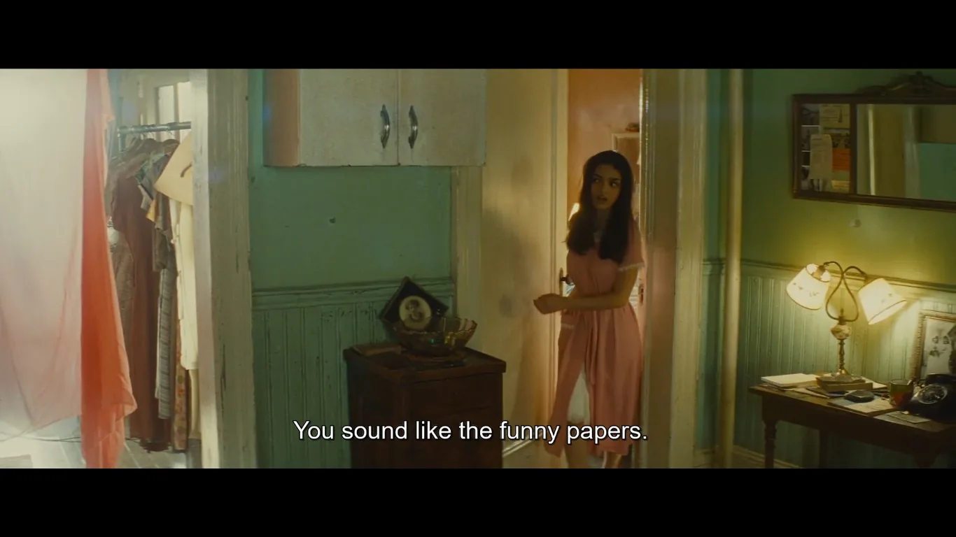 The scene takes place in a cozy, dimly lit room, giving off a warm and nostalgic atmosphere. A young woman, wearing a pink nightgown, stands in a doorway. Her expression is slightly surprised or curious, as if responding to someone unseen in another room. The walls are painted a soft green, with wooden wainscoting adding a vintage touch. A small triangular shelf holds a framed photo and a decorative bowl, hinting at a personal and lived-in space.
To the left, a clothes rack with various garments is partially visible, suggesting this might be a bedroom or dressing area. On the right, a desk holds a table lamp with two shades, papers, and a vintage rotary phone, emphasizing the period setting. The lighting is soft and yellow, contributing to the overall intimate and historical feel of the scene. The caption at the bottom reads, "You sound like the funny papers," indicating a casual or humorous exchange.