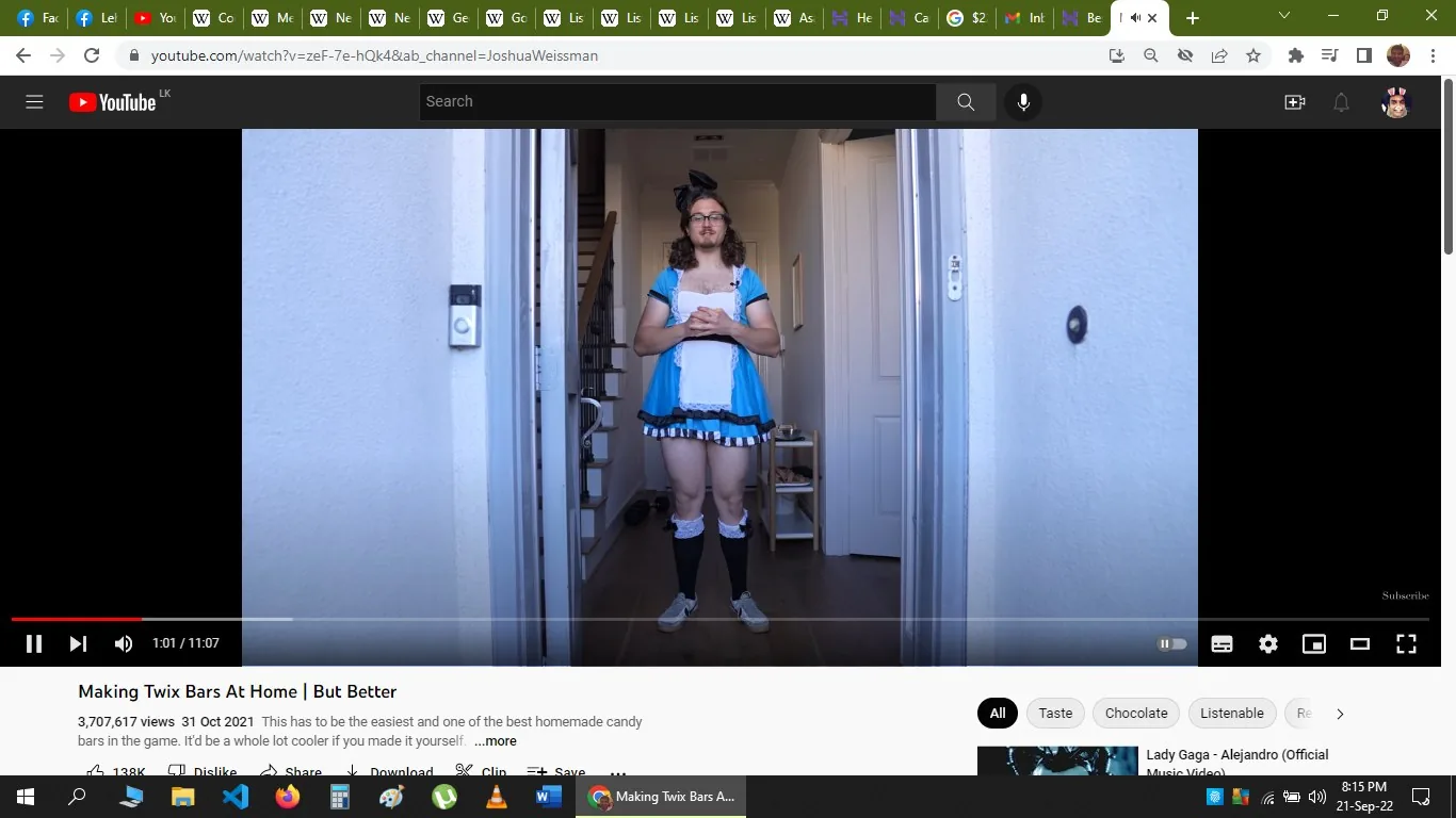 The image is a YouTube screenshot showing Joshua Weissman in a humorous and unconventional outfit, standing in an open doorway. He is dressed in a blue and white maid costume, complete with a frilly skirt, an apron, and knee-high socks. His expression is neutral, and he stands with his hands clasped in front of him. The setting is a residential interior, with a staircase and some household items visible in the background. The lighting is bright and natural, contributing to a casual and playful atmosphere. This scene is part of a video titled "Making Twix Bars At Home | But Better," which is hosted on Joshua Weissman's YouTube channel. The video, with over 3.7 million views, appears to focus on cooking, specifically making homemade Twix bars. The layout of the YouTube interface is visible, including buttons for liking, sharing, and more.