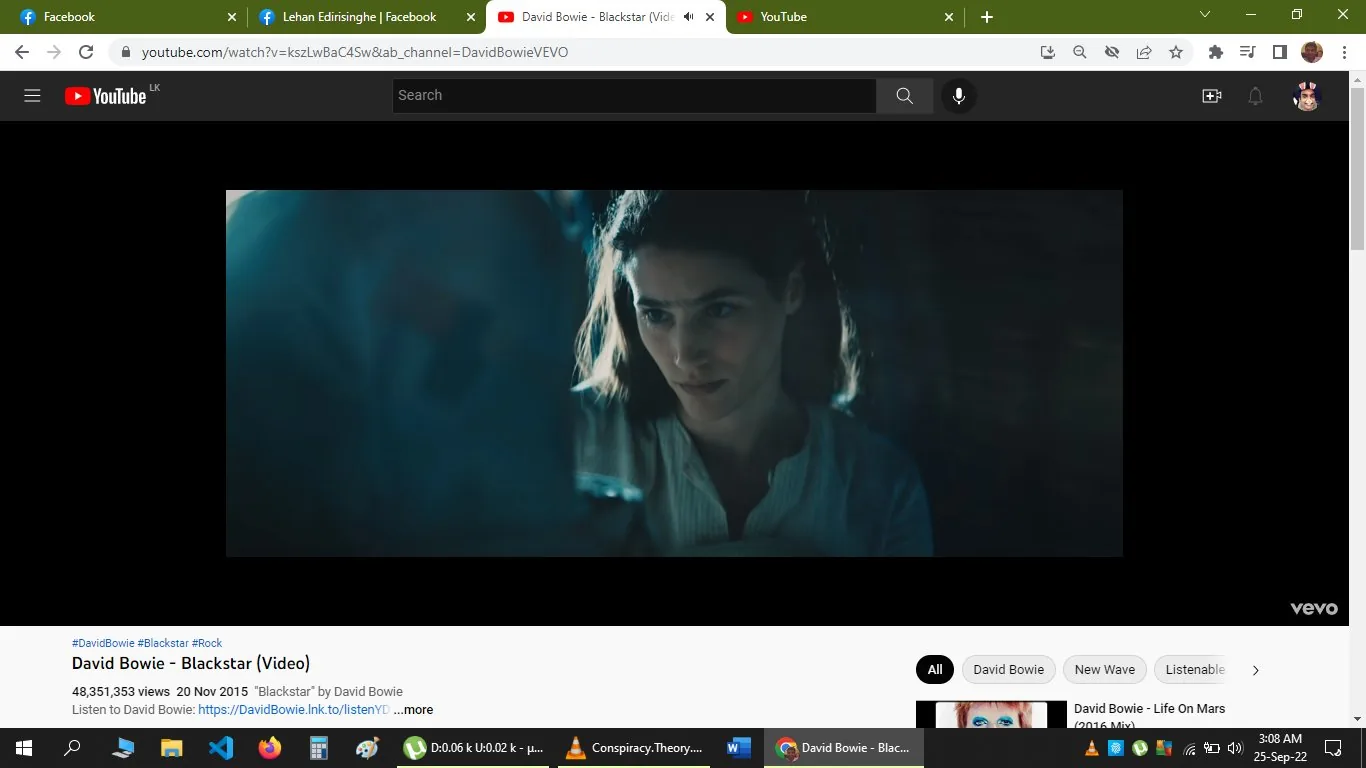 The image is a screenshot from a YouTube video titled "David Bowie - Blackstar (Video)" on the DavidBowieVEVO channel. It depicts a scene from the music video for the song "Blackstar" by David Bowie. The setting is dimly lit, creating a mysterious and moody atmosphere. A woman is prominently featured in the foreground, looking intently at something off-screen. Her expression is serious and contemplative, adding to the video's enigmatic tone. The lighting casts a bluish tint, enhancing the otherworldly feel. The woman's attire is simple, consisting of a light-colored top, which contrasts with the darker, shadowy background. The screenshot includes visible YouTube interface elements, such as the video title, view count, and other metadata. Above the video, browser tabs are open, indicating other sites like Facebook and additional YouTube tabs. The video is produced by VEVO, and topic tags include #DavidBowie, #Blackstar, and #Rock.
- Title: "David Bowie - Blackstar (Video)"
- Channel / profile: "DavidBowieVEVO"
- Site / app: "YouTube"