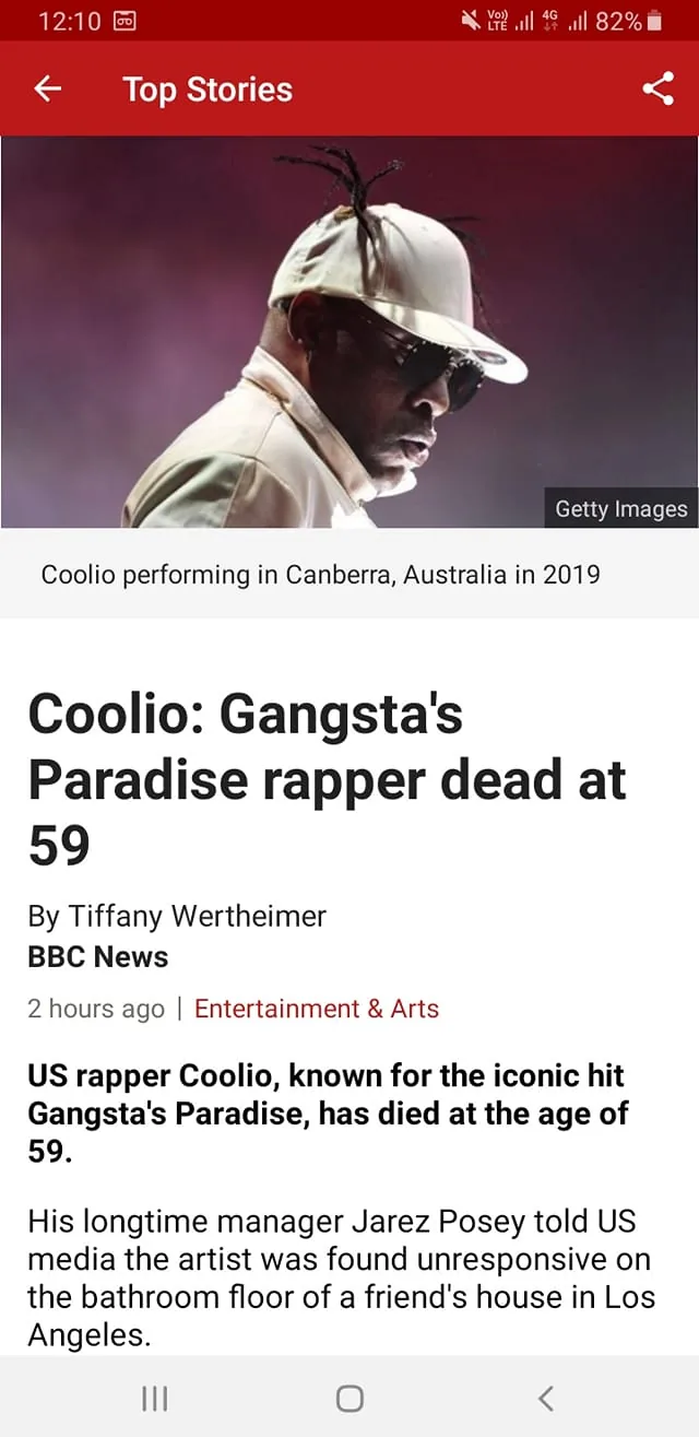 The image is a screenshot from the BBC News app featuring an article about the death of rapper Coolio. The visual component shows a photograph of Coolio performing on stage in Canberra, Australia, in 2019. He is wearing a white cap with his distinctive hairstyle protruding, and he is dressed in a white outfit. The lighting creates a dramatic effect with a spotlight focusing on him, highlighting his profile against a dark background. The article headline reads, "Coolio: Gangsta's Paradise rapper dead at 59," followed by the author's name, Tiffany Wertheimer, and the publisher, BBC News. The text states that Coolio, known for his hit "Gangsta's Paradise," has passed away at the age of 59. It further mentions a statement from his manager, Jarez Posey, about the circumstances of Coolio's passing. The interface shows that this is part of the "Top Stories" section, emphasizing its importance in the news cycle.