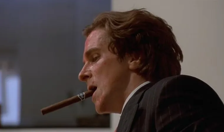 ```
Christian Bale as Patrick Bateman is depicted in a scene from the film "American Psycho." The image captures a close-up of Bateman's face, turned slightly to the side, highlighting his intense expression. He is smoking a cigar, which adds to the character's portrayal of wealth and power. His hair is neatly styled, and he is dressed in a dark pinstripe suit and white shirt, typical of his character's affluent lifestyle. The lighting is dim, casting shadows that enhance the dramatic and menacing mood. The background is blurred, focusing the viewer's attention on Bateman's face and the cigar. The overall composition conveys a sense of tension and underlying aggression, characteristic of the film's tone.