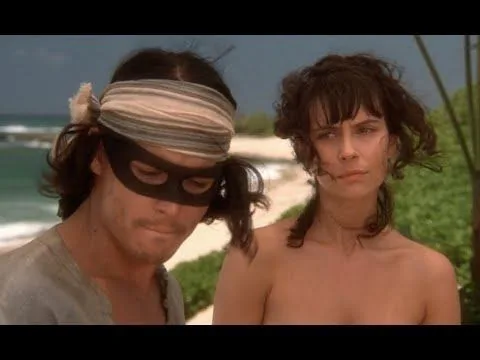 In this scene from the film "Don Juan DeMarco," Johnny Depp is prominently featured. He wears a distinctive black eye mask and a striped headscarf tied around his long hair, capturing the romantic and adventurous spirit of his character. Beside him, a woman with dark, curly hair gazes ahead, her expression suggesting contemplation or curiosity. The background reveals a picturesque beach setting with the ocean and a clear blue sky, enhancing the romantic and adventurous atmosphere typical of the film. The lighting is natural and bright, emphasizing the daytime setting. Johnny Depp's character is dressed in a casual, slightly rugged style, fitting the adventurous theme, while the woman's attire is minimal, focusing attention on her facial expression and interaction with Depp. The composition places both characters in the foreground, with the serene beach landscape adding depth to the scene.