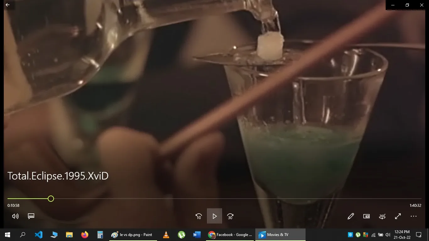 The image is a close-up screenshot taken from what appears to be a scene in the film "Total Eclipse." The focus is on a glass partially filled with a bluish liquid. In the foreground, a hand is holding a bottle, pouring a clear liquid over a sugar cube placed on what seems to be an absinthe spoon resting on the glass. The scene is likely depicting the preparation of an absinthe drink, a process traditionally involving dripping water over a sugar cube into the absinthe. The lighting is warm and dim, creating an intimate and old-fashioned atmosphere typical of 19th-century settings. The background is blurred, emphasizing the action in the foreground. The visible media player interface at the bottom of the image shows the title "Total.Eclipse.1995.XviD," indicating that this is a still from the movie "Total Eclipse," which is being viewed using the Movies & TV app.