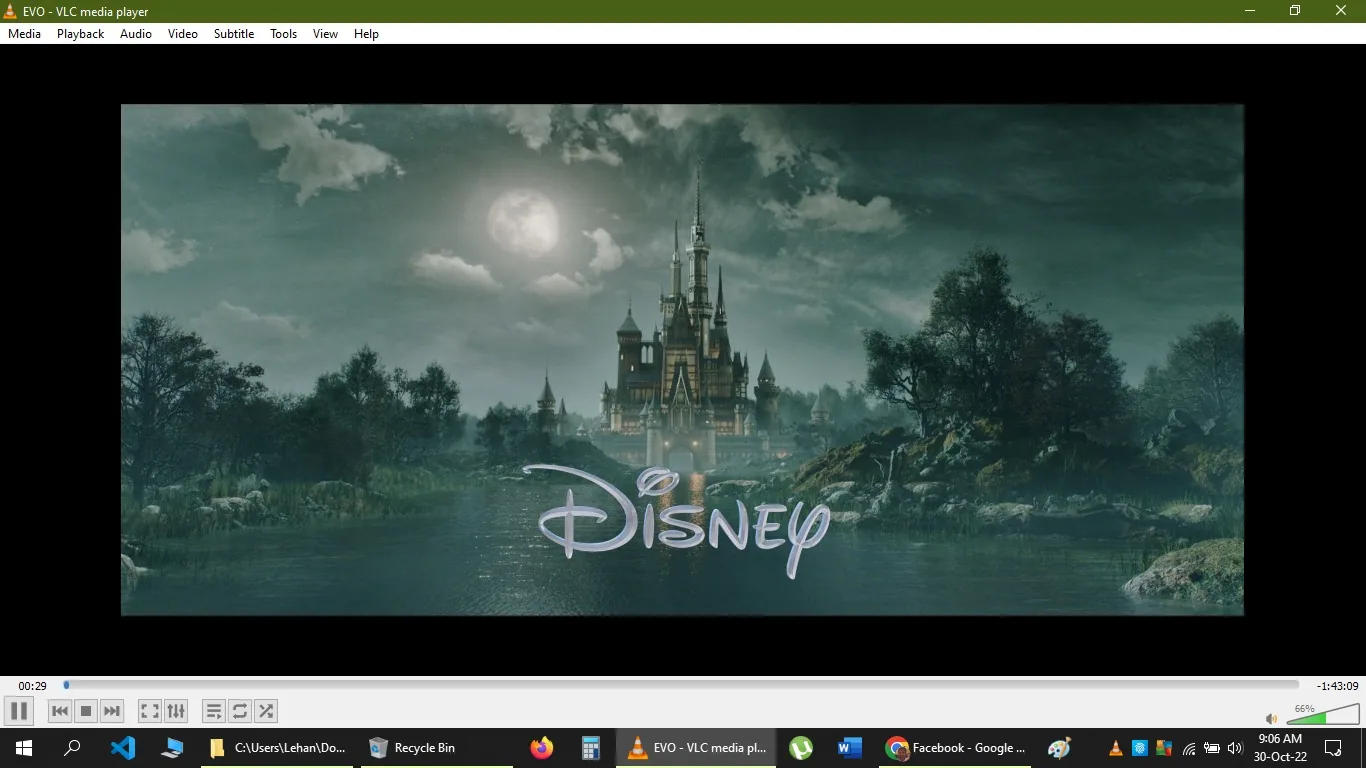 The image is a screenshot from VLC media player, showcasing an iconic Disney castle under a moonlit sky. This classic Disney introduction features a castle set amid a serene landscape, surrounded by lush trees and a calm body of water reflecting the castle's lights. The sky is overcast with dense clouds, and the full moon casts a subtle glow over the scene, enhancing the magical and fantastical atmosphere typical of Disney productions. The Disney logo is prominently displayed in the foreground, written in its characteristic, whimsical font. The architectural style of the castle is grand and fairy-tale-like, with multiple spires and towers. The overall tone of the image is mystical, capturing the enchanting essence Disney is known for. The screenshot is taken from the VLC media player interface, which is visible with media controls and a green-themed menu bar at the top.