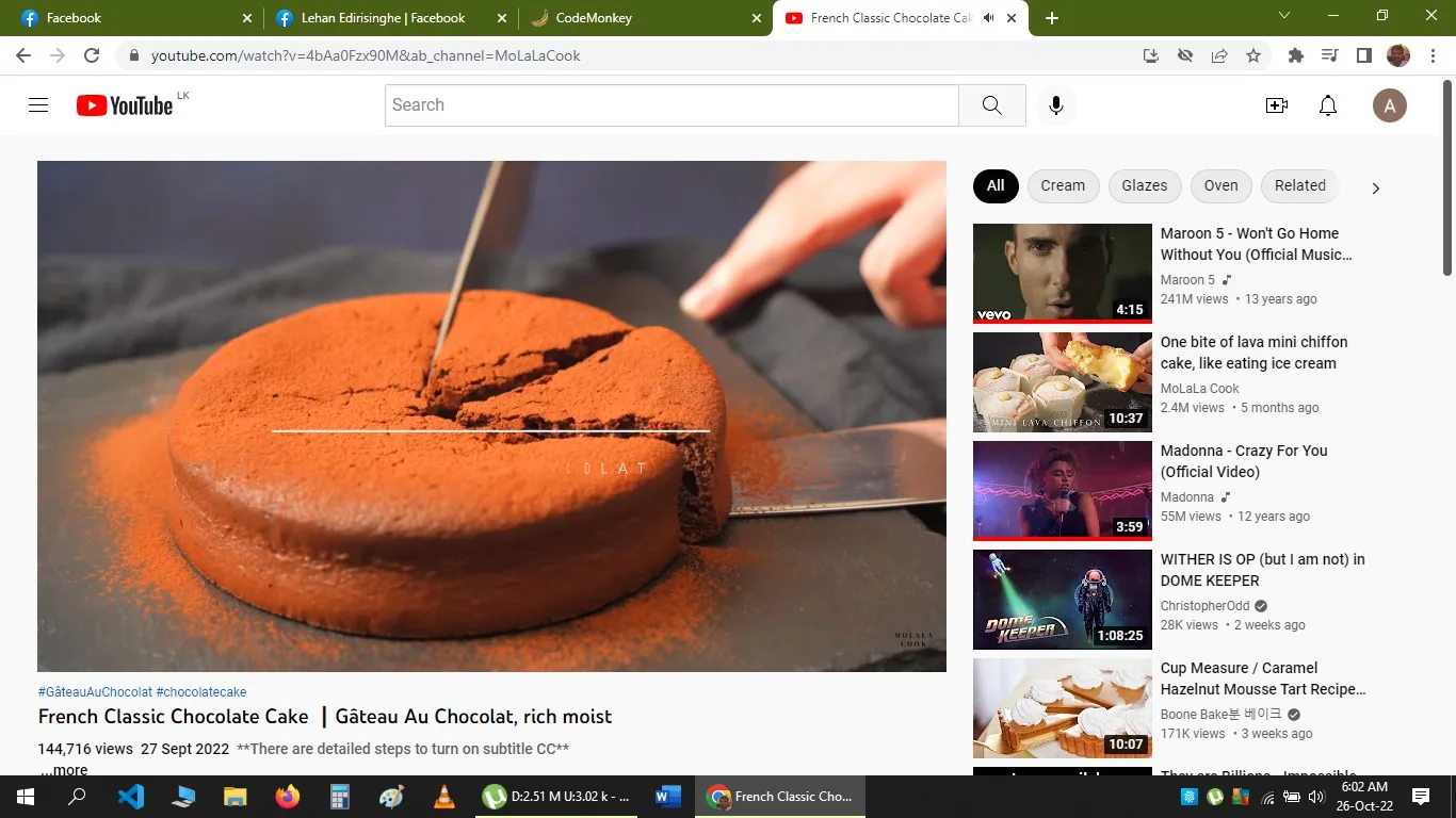 This image is a screenshot from a YouTube video, displaying the video page of a cooking tutorial titled "French Classic Chocolate Cake | Gâteau Au Chocolat, rich moist" by the channel MoLaLaCook. The central image shows a close-up of a rich, chocolate cake being sliced, with a dusting of cocoa powder on top. A hand is using a knife to cut a slice from the cake, which is placed on a dark surface, likely a countertop or cutting board, highlighting the deep brown color and appealing texture of the cake.
The right side of the screenshot includes a list of related video thumbnails, featuring a mix of music videos and other cooking tutorials. The page header shows the YouTube logo and standard interface elements, while the search bar and user profile icon are visible at the top. The video's view count and publication date, "144,716 views 27 Sept 2022," are displayed under the video title. The hashtags "#GâteauAuChocolat" and "#chocolatecake" suggest the video is tagged for users interested in those topics.
The composition emphasizes the cake's texture and richness, making it visually appealing for viewers interested in baking. The overall aesthetic is clean and inviting, typical of professional cooking channels aiming to teach and inspire.