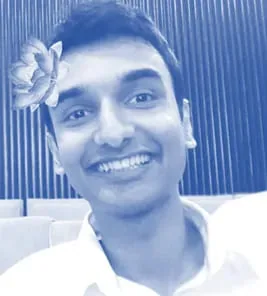 Lehan Edirisinghe is depicted in this image, which has been tinted in blue, giving it a monochromatic effect. The setting appears to be indoors, possibly in a theater or lecture hall, suggested by the rows of upholstered seats in the background. Lehan is smiling broadly, creating a joyful and approachable impression. His attire is a white shirt, which contrasts with the blue overlay. A digital effect adds a flower to his hair, giving a playful and whimsical touch to the image. The background features vertical wooden slats, adding texture and structure to the scene. The lighting is even, highlighting his facial features, and the overall mood of the image is lighthearted and cheerful.
