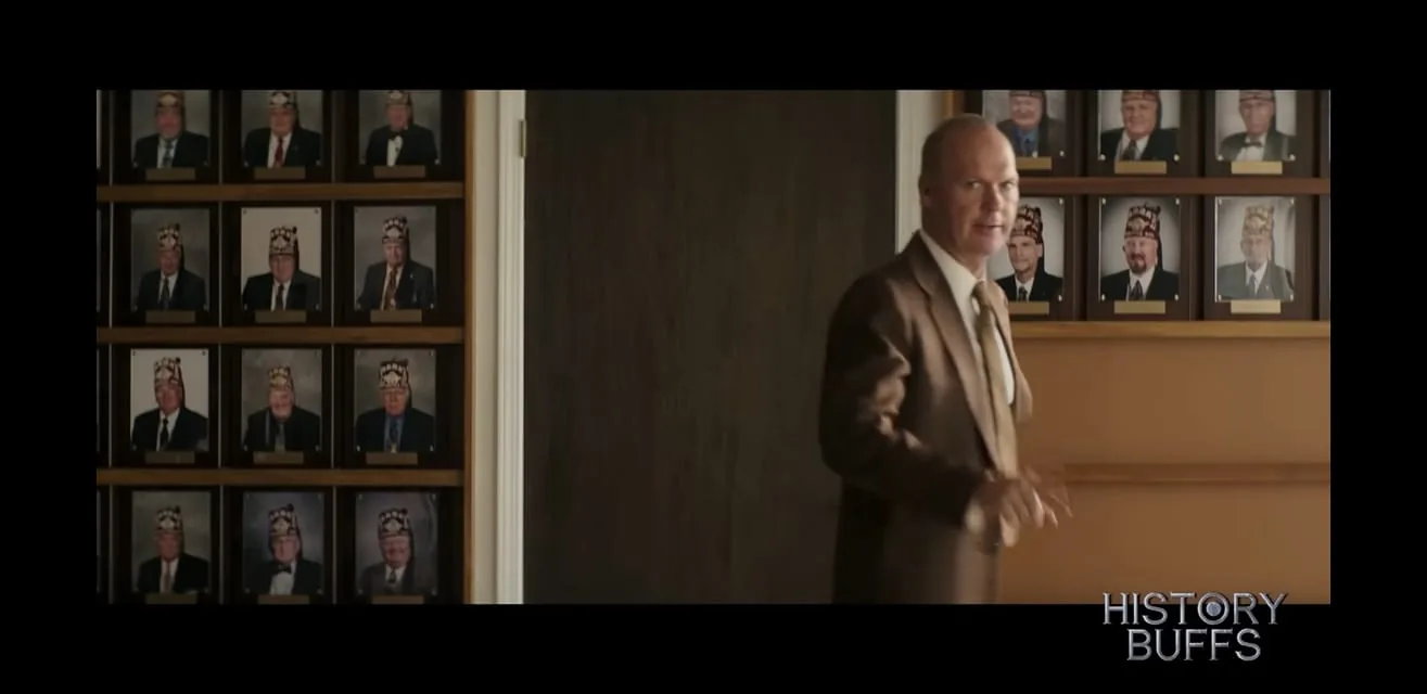 The image is a frame from a video on the YouTube channel "HISTORY BUFFS." The scene is set indoors and features a man in a brown suit, standing in front of a wall displaying numerous framed portraits. The portraits show individuals wearing hats that resemble those worn by members of fraternal organizations. Each portrait has a nameplate beneath it, though the names are not legible. The man in the foreground appears to be in motion, possibly speaking or gesturing toward the viewer or someone off-screen. The lighting is soft, providing a warm and formal atmosphere to the room. The channel "HISTORY BUFFS" is known for analyzing films and discussing their historical accuracy. The video frame does not include any visible text outside of the channel's watermark.