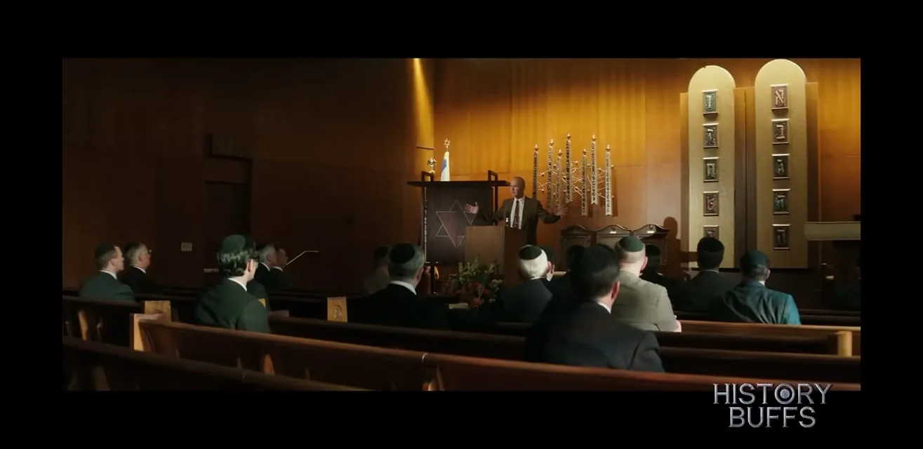 This image depicts a scene set in a synagogue, where a man is standing at a podium speaking to an audience. The setting gives a feeling of solemnity, with rows of wooden pews occupied by men wearing dark suits and kippahs. The speaker, positioned at the front, is gesturing with open arms, suggesting he is engaged in delivering a speech or sermon. Behind him, the wall is adorned with religious symbols, including a large Star of David on the podium and decorative elements related to Jewish tradition. The lighting is warm and focused, emphasizing the speaker and creating a calm, respectful atmosphere. In the lower right corner, the text "HISTORY BUFFS" is visible, indicating the channel or profile where this content is featured. The presence of such specific religious iconography and attire suggests this scene might be part of a documentary or historical discussion about Jewish culture or history.