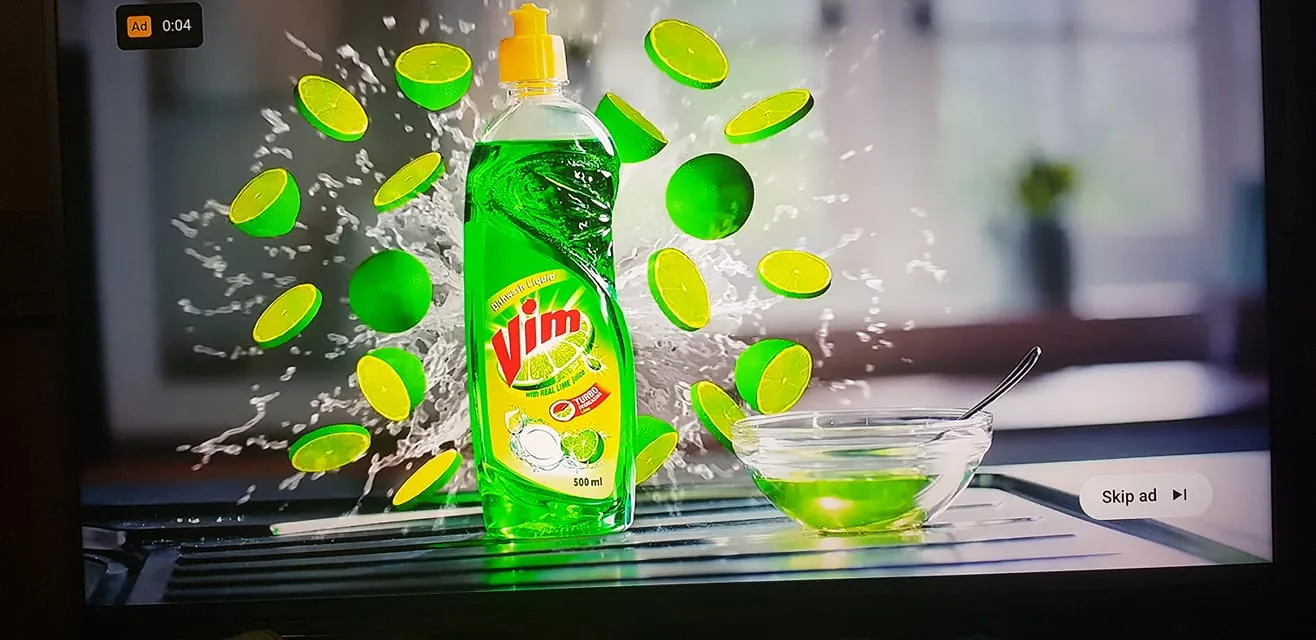 The image is a vibrant advertisement for a dishwashing liquid product named Vim. The central focus is a bottle of Vim dishwashing liquid, prominently displaying its bright green color and clear plastic design. The label on the bottle features the brand name "Vim" in bold red letters, accompanied by the phrase "With real lime juice" and the word "Turbo," suggesting a powerful cleaning effect. Surrounding the bottle are numerous lime slices and whole limes, dynamically frozen in mid-air, with a splash effect adding a sense of freshness and vitality. In front of the bottle sits a small glass bowl filled with green liquid, echoing the product's color. The overall setting appears to be a kitchen, with a blurred background that suggests a modern, clean environment, enhancing the product's appeal for home use. This composition, with its energetic and clean visual elements, effectively highlights the product's citrus freshness and cleaning prowess.