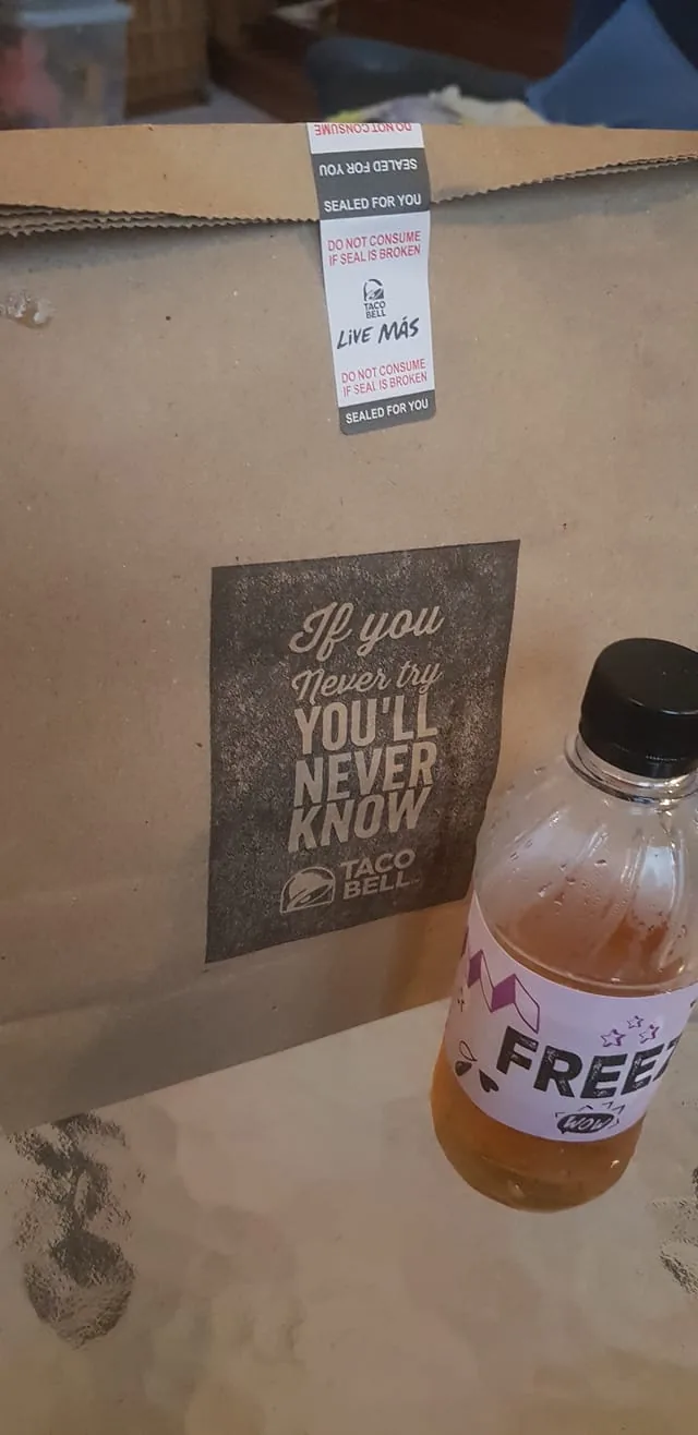 The image shows a Taco Bell paper bag with visible branding and a motivational quote printed on it. The text on the bag reads, "If you never try you'll never know," followed by the Taco Bell logo. The bag is sealed with a sticker that says, "Do not consume if seal is broken," emphasizing food safety. In the foreground, there is a small plastic bottle filled with a light-colored beverage. The bottle has a label with the word "FREEZE" and various decorative elements, suggesting it's a frozen drink from Taco Bell. The scene appears to be indoors, possibly at a home or casual setting. The background is slightly blurred, suggesting the focus is on the bag and the drink. The overall lighting is warm and natural, likely from ambient indoor light.