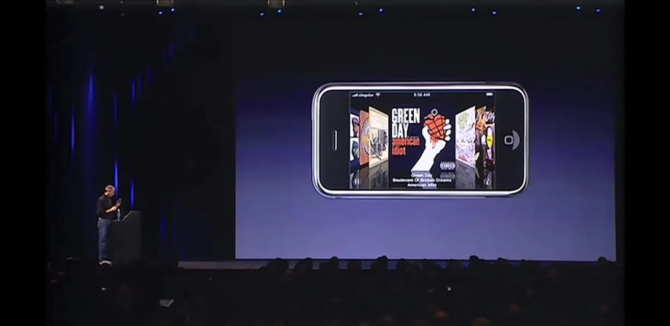 Steve Jobs is on stage during a presentation, standing next to a podium with a large screen behind him. The screen prominently features an image of a smartphone displaying a cover flow of music albums. The album "American Idiot" by Green Day is visible, alongside others. The setting is a dimly lit auditorium, likely filled with an audience, as indicated by the silhouettes below the stage. Steve Jobs is casually dressed in his signature black turtleneck shirt and jeans. The lighting focuses on the stage, highlighting Jobs and the large screen. The phone display showcases the album art in a horizontal orientation, suggesting a feature related to music library navigation on the device. This scene captures a moment in a technological announcement, emphasizing the capacity for media storage and playback on a new smartphone.