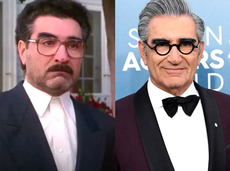 ```
The image shows a side-by-side comparison of a famous actor, known from the movie "American Pie," highlighting his transformation over the years. On the left, the actor appears younger, with a fuller mustache and hair, wearing large, square glasses and a dark suit. His expression is serious, and the background features a blurred outdoor setting, suggesting a scene from a film.
On the right, the actor is older and dressed formally for an event, likely an award show. He sports a tuxedo with a bow tie and has stylish, round glasses. His hair is neatly styled, and he has a more relaxed, confident expression. The background is a blue step-and-repeat banner, indicating a formal or red carpet setting. The contrast in his appearance highlights both aging and a change in style over time.