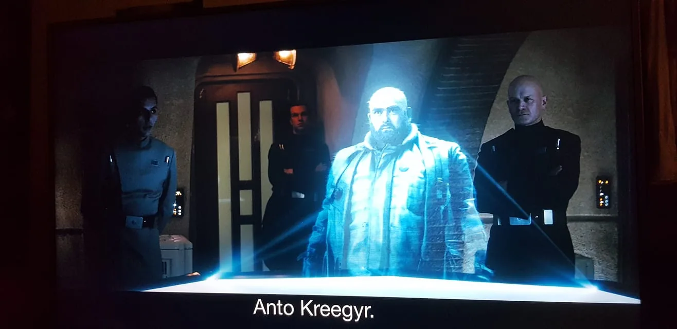 The image is a screenshot from a scene depicting an indoor setting with dim lighting, suggesting a tense or serious atmosphere. In the center of the frame, there is a holographic projection of a character named Anto Kreegyr, who appears transparent with a bluish glow typical of holograms in science fiction settings. He stands confidently, with a sturdy build, wearing a jacket that adds to his commanding presence.
Surrounding the hologram are three individuals dressed in dark military-style uniforms, suggesting they are part of a formal or authoritative organization. Their postures are rigid and attentive, indicating a possible interrogation or briefing scenario. The background features a door with a futuristic design, enhancing the sci-fi theme. The overall lighting is muted, with the hologram being the main source of illumination, creating stark contrasts and shadows that emphasize the gravity of the scene. The caption at the bottom reads "Anto Kreegyr," identifying the holographic character.