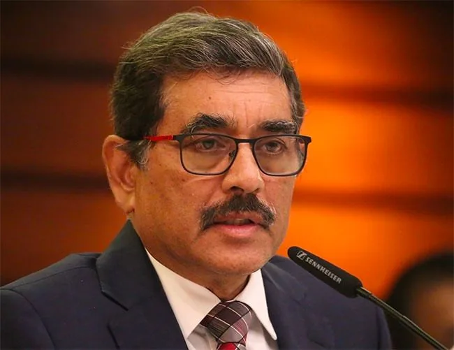 Nandalal Weerasinghe is depicted in this image, speaking at a conference or formal event. He is positioned in front of a microphone branded with "Sennheiser," suggesting a professional setting. Weerasinghe is dressed in a formal suit and tie, featuring a dark jacket and a patterned tie that complements his attire. His expression is attentive and focused, indicative of someone addressing an audience or participating in a discussion. The background is softly blurred with warm tones, enhancing the subject’s prominence in the image. Lighting is well-balanced, highlighting his features and the crispness of his attire. His glasses have a subtle red detail on the temples, adding a touch of color to his classic look. The overall composition and lighting suggest a serious and professional atmosphere, typical of a corporate or academic setting.