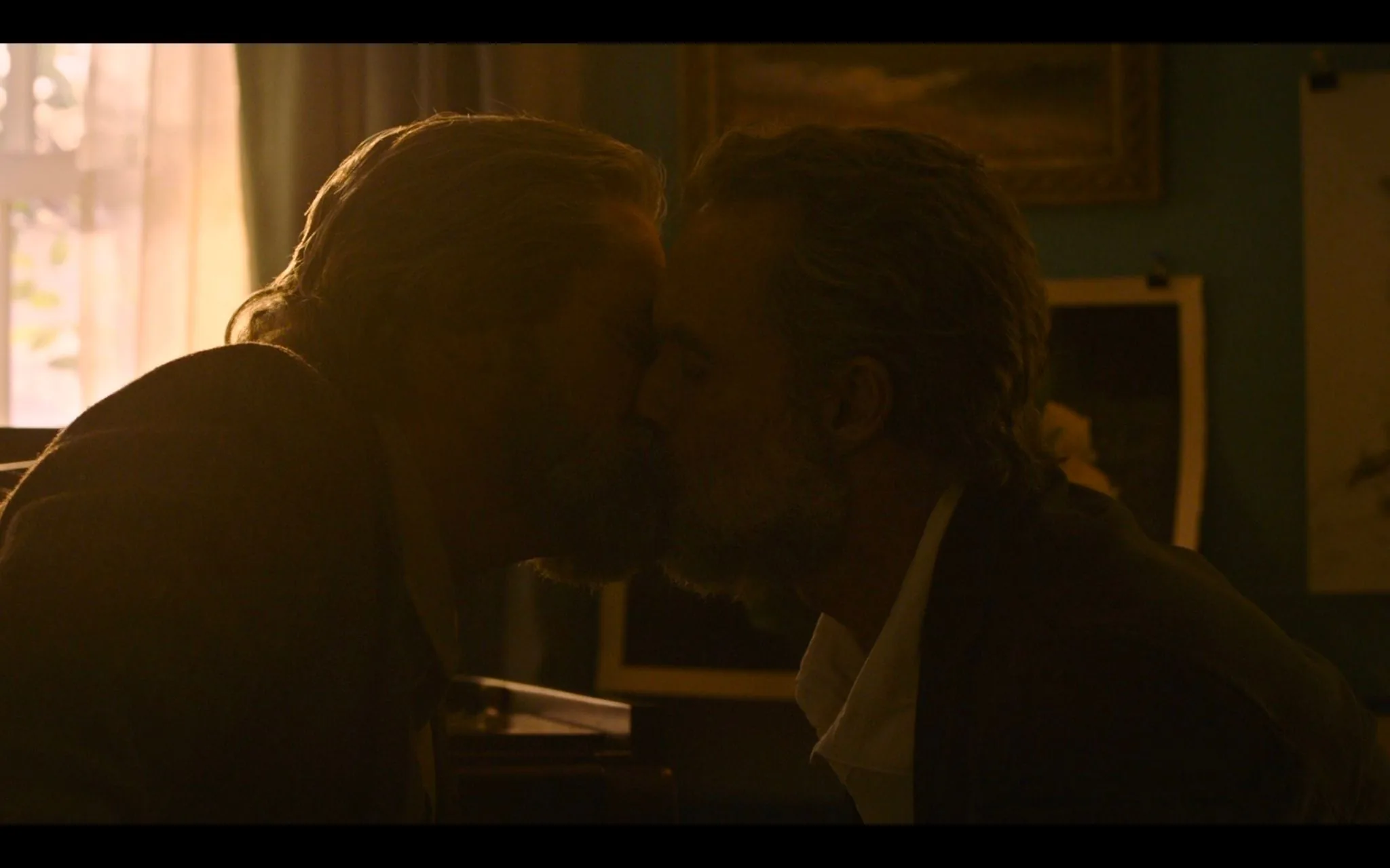 In this screenshot from "The Last of Us," there is an intimate and emotional scene featuring Nick Offerman. Two men are leaning in for a kiss, suggesting a moment of deep affection and connection. The lighting is warm and dim, casting a gentle glow that emphasizes the tenderness of the scene. The background shows a softly lit room with art and possibly photographs on the walls, adding a personal and cozy atmosphere.
The characters are seated closely, suggesting familiarity and comfort with one another. The attire is casual yet slightly formal, with one wearing a light shirt and the other in a darker outfit, which contrasts softly with the room's muted tones. The focus is on their expressions and the closeness of their gesture, conveying a sense of vulnerability and love. The overall mood is somber yet heartfelt, fitting the dramatic and narrative-driven nature of "The Last of Us."