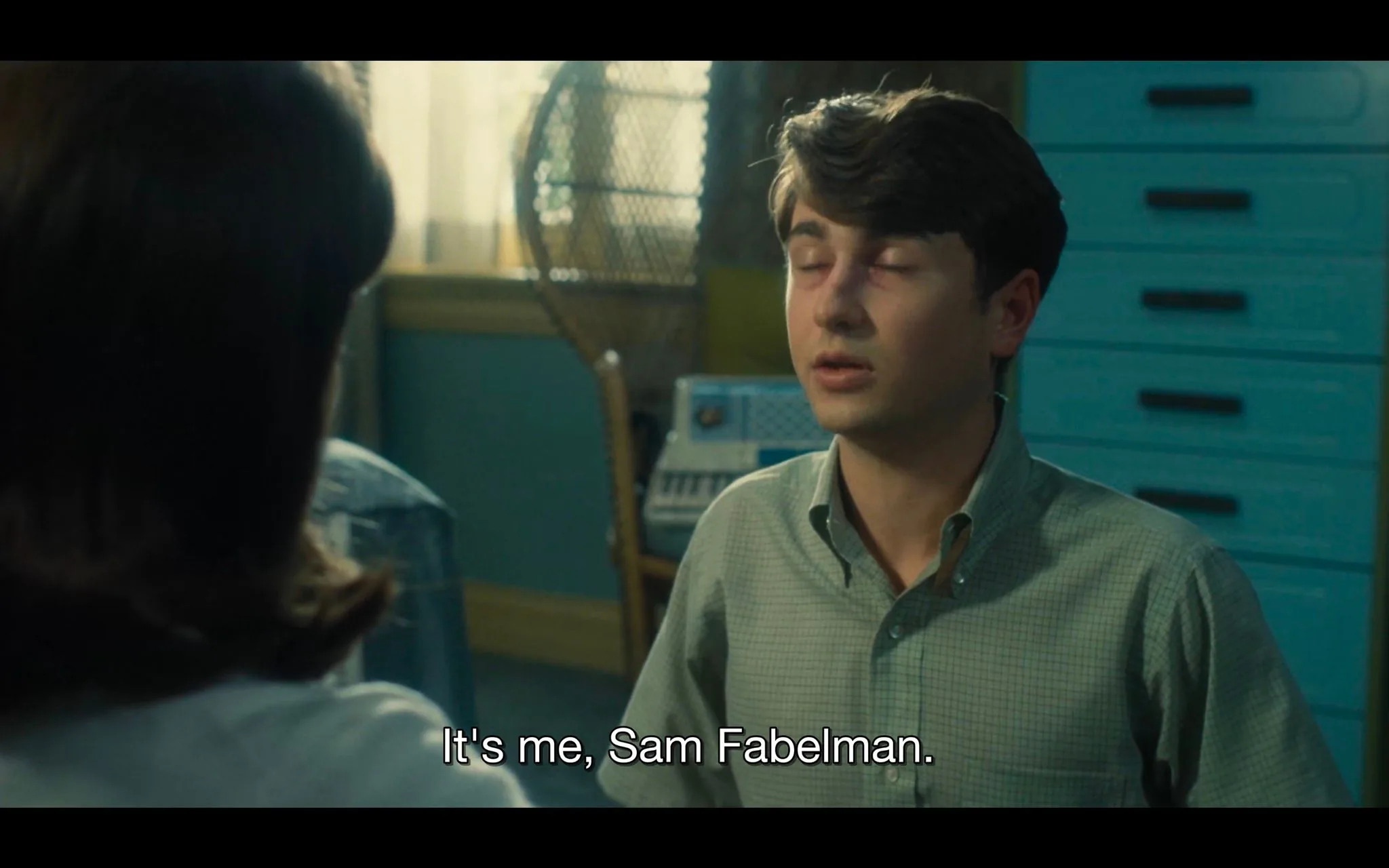 ```
In this scene from "The Fabelmans," a young man identified as Sam Fabelman is shown sitting indoors. The lighting is warm, suggesting a domestic setting. The focus is on Sam, who has a serious expression and a visible bruise under his eye, indicating he might have been in a recent altercation. He is wearing a light plaid shirt, which complements the casual and personal atmosphere of the scene. In the background, there's a wicker chair and a piece of electronic equipment, possibly a typewriter or radio, adding to the mid-20th century ambiance. The furniture and decor, including a blue cabinet, reinforce the period setting. The over-the-shoulder shot framing implies a dialogue with another character, whose back is partially visible. The caption reads, "It's me, Sam Fabelman," emphasizing his identity and possibly a significant moment in the narrative. The scene captures a moment of introspection or revelation for Sam.