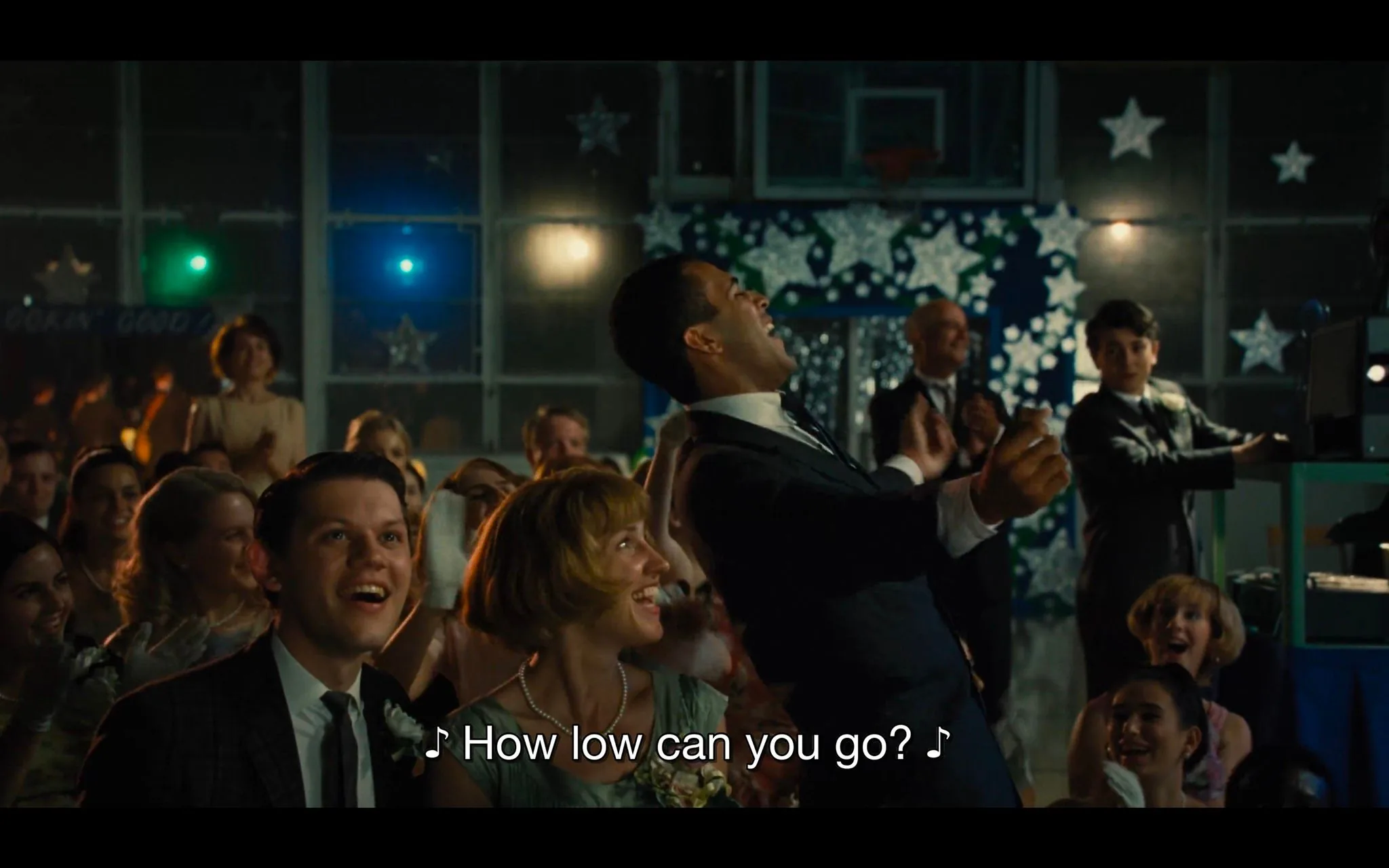 ```
1) Rich description: The scene captures a lively indoor event, possibly a dance or a celebration, with a crowd of people enjoying the moment. The setting is decorated with star-shaped cutouts on the walls, suggesting a festive or themed occasion. In the foreground, a man in a dark suit and tie is leaning back enthusiastically, appearing to dance or participate in a game. He seems to be enjoying himself, with a broad smile and animated posture. Nearby, a woman with short blonde hair, wearing a green dress and pearls, looks on with delight. The crowd around them, dressed in semi-formal attire, is engaged and smiling, adding to the energetic atmosphere. The lighting is warm, highlighting the cheerful expressions of the attendees. In the background, a young person in a suit appears to be operating equipment, possibly controlling music or lights. The caption reads, "♪ How low can you go? ♪," indicating the soundtrack or activity involves a limbo dance or similar challenge.