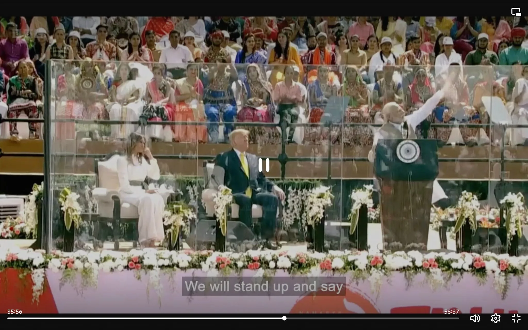 The image captures an outdoor event scene featuring Melania, Trump, and Modi. They are positioned on a stage with a large audience in the background, seated in a stadium-like setting. The stage is adorned with an array of flowers, adding vibrant colors to the setting. A glass barrier separates the stage from the audience.
On the right side of the stage, Modi is at a podium, gesturing energetically, likely delivering a speech. He wears a white kurta and a dark vest, characteristic of his typical attire. In the center, Trump is seated, wearing a dark suit with a yellow tie, appearing attentive to the speech. Next to him, Melania sits elegantly in a white outfit, with a thoughtful expression.
The audience, visible behind the glass, features people in colorful traditional attire, indicating a culturally diverse gathering. The lighting is bright, suggesting a daytime event. The caption at the bottom of the image reads, "We will stand up and say," reflecting the speech's motivational tone.