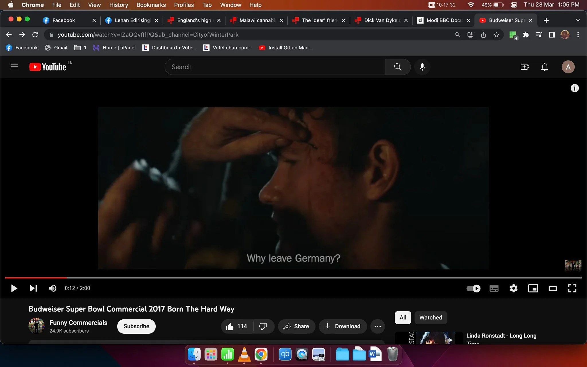 The image is a video frame from a YouTube video titled "Budweiser Super Bowl Commercial 2017 Born The Hard Way," uploaded by the channel "Funny Commercials." The scene depicted is a close-up shot of a person, suggesting an emotional moment, with another hand gently touching their forehead. The lighting is dim, creating a dramatic and intense atmosphere. The background is blurred, focusing attention on the subject's face. The video features subtitles with the line “Why leave Germany?” indicating a narrative about leaving or migration, possibly reflecting the historical journey of Budweiser’s founders. The video is part of a Super Bowl commercial, known for storytelling and cinematic quality. The composition uses dark tones, enhancing the emotional depth and seriousness of the scene.