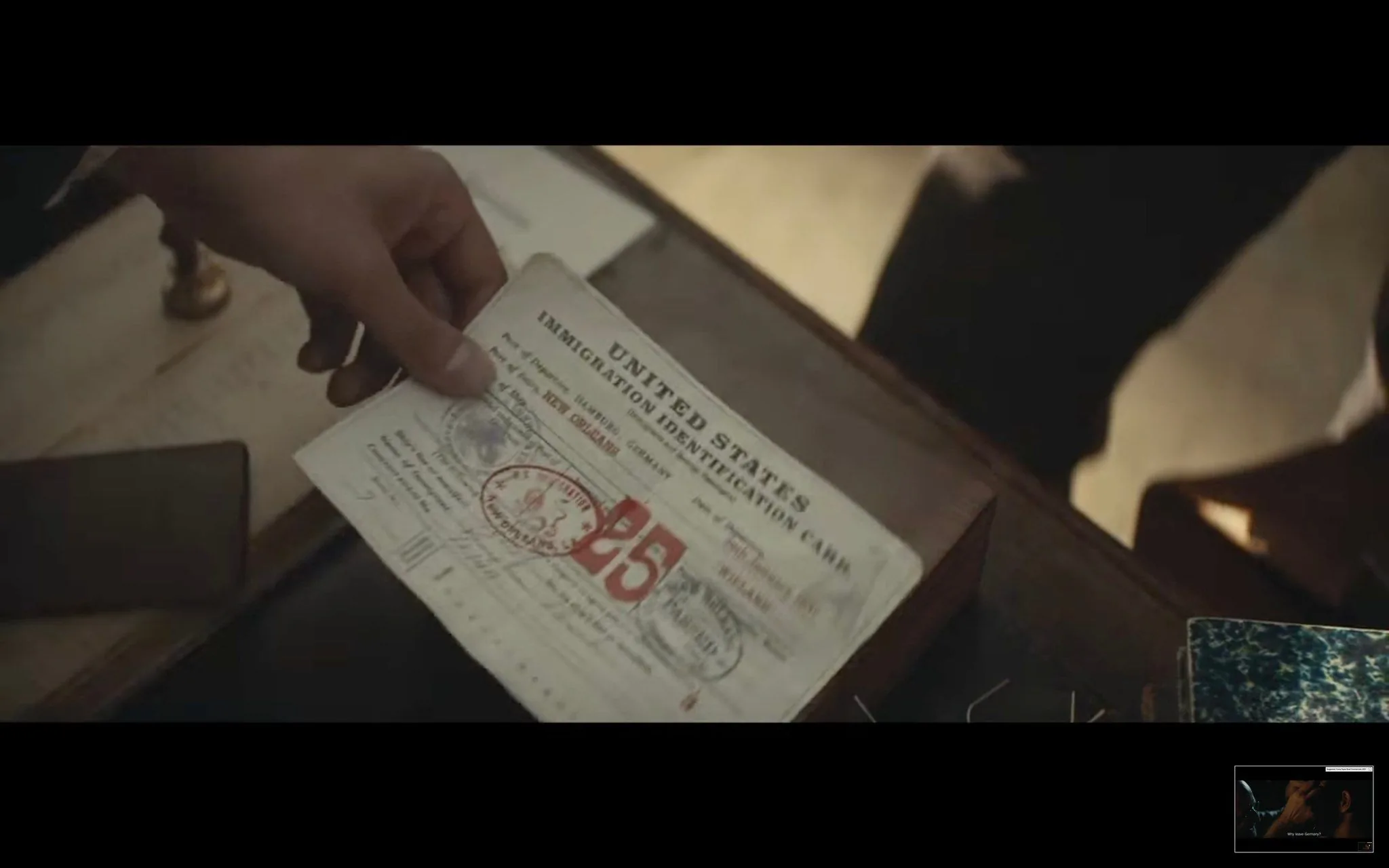 The image shows a scene likely from a film or TV series, capturing a close-up of a hand holding an immigration identification card. The document is labeled "UNITED STATES IMMIGRATION IDENTIFICATION CARD" at the top. Additional visible text includes "Port of New York" and a large "25" along with other markings and stamps, suggesting it is an official document related to immigration or travel. The hand holding the card is positioned over a desk that appears to be cluttered with papers and a notebook, indicating a typical office or administrative setting. The lighting is soft, creating a warm ambiance that adds a sense of nostalgia or historical context to the scene. This gives the impression of a period piece, possibly set in the early to mid-20th century, as suggested by the style of the document and the muted color palette. The focus on the card implies its importance within the narrative, possibly serving as a plot device related to identity, travel, or legal status.