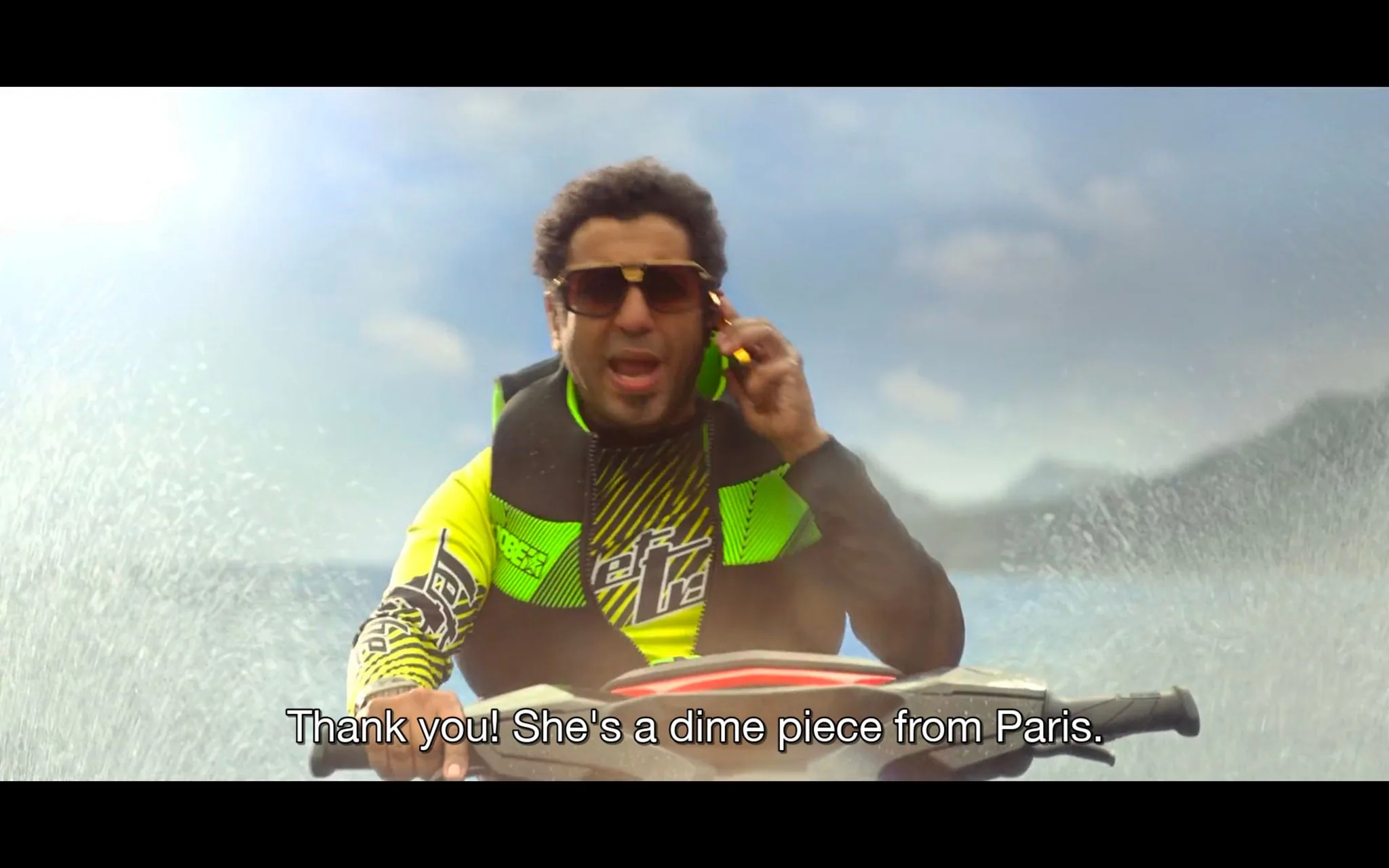In this scene, a man is riding a jet ski on a body of water, captured in bright, sunny lighting that highlights the vibrant colors of his outfit. He is wearing a neon yellow and black life vest with matching attire that features bold patterns. The man sports large sunglasses and is talking on a mobile phone held to his ear, suggesting a lively or animated conversation. The background shows a clear blue sky with hints of white clouds, and the water is splashing around, emphasizing the action and movement. The scene is energetic, with the man appearing focused and excited. The subtitles indicate he's talking about someone from Paris, using colloquial language. The visual style is dynamic, capturing the essence of outdoor adventure and communication on the go.
- Captions / subtitles: "Thank you! She's a dime piece from Paris."
