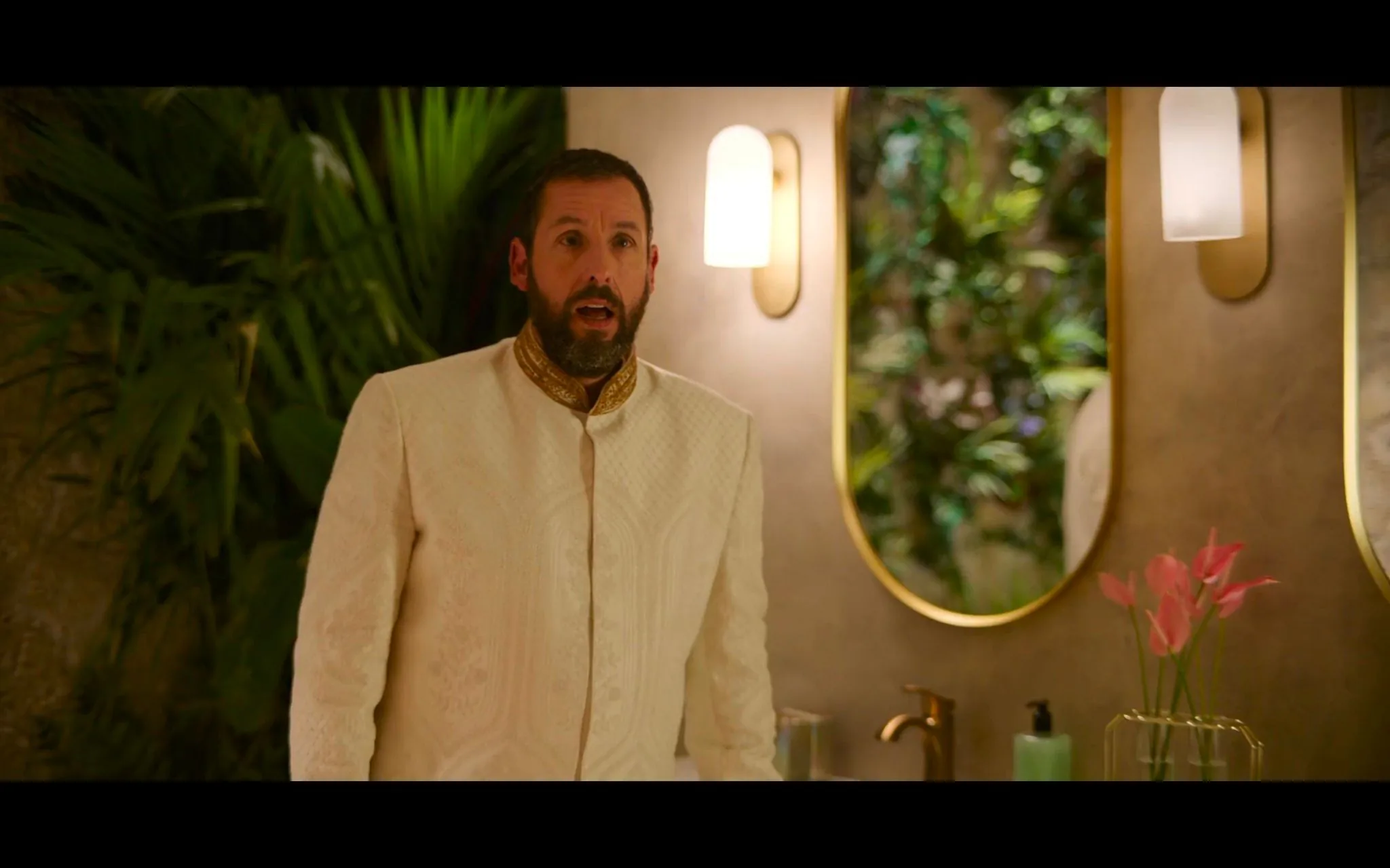 Adam Sandler is depicted in a luxurious bathroom setting, wearing ornate Indian attire. His outfit is a sophisticated cream-colored sherwani with intricate embroidery, featuring a gold collar that adds an elegant touch. Sandler's facial expression appears surprised or engaged, with his mouth slightly open as if speaking or reacting to something outside the frame. The room is warmly lit, creating a cozy and inviting atmosphere.
In the background, a large mirror reflects a wall with lush green plants, adding a natural element to the space. The bathroom is tastefully decorated, featuring minimalist design elements with two wall-mounted lights beside the mirror. A vase with pink flowers is placed on the countertop, alongside a green soap dispenser and a gold-colored faucet, contributing to the overall aesthetic of understated luxury. The image conveys a sense of opulence and style, with Adam Sandler's presence adding a focal point to the scene.