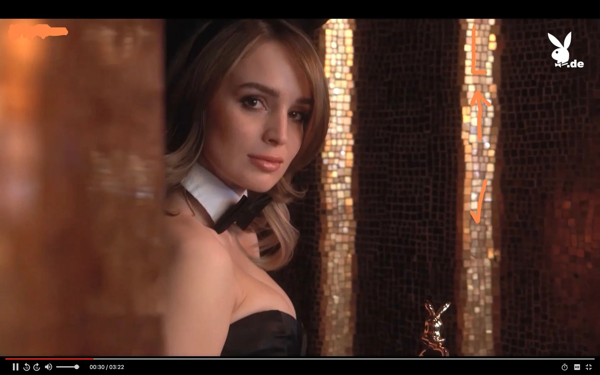 Julia Rommelt is captured in an elegant indoor setting, exuding a sense of glamour and allure. The image features her in a close-up, focused on her face and upper body. She is dressed in a classic Playboy Bunny outfit, which includes a black strapless dress, a white collar, and a black bow tie. Her hair is styled in soft waves, complementing the sophisticated look. The background reveals a shimmering mosaic wall that adds a touch of luxury and depth to the scene. The lighting is warm, highlighting her features and the glossy texture of the wall tiles. In the bottom right corner, a small metallic Playboy bunny emblem is visible, reinforcing the brand identity. The image conveys elegance and poise, typical of high-fashion photography associated with the Playboy brand. The presence of ".de" hints at German-language content, possibly indicating a regional edition or version of Playboy.