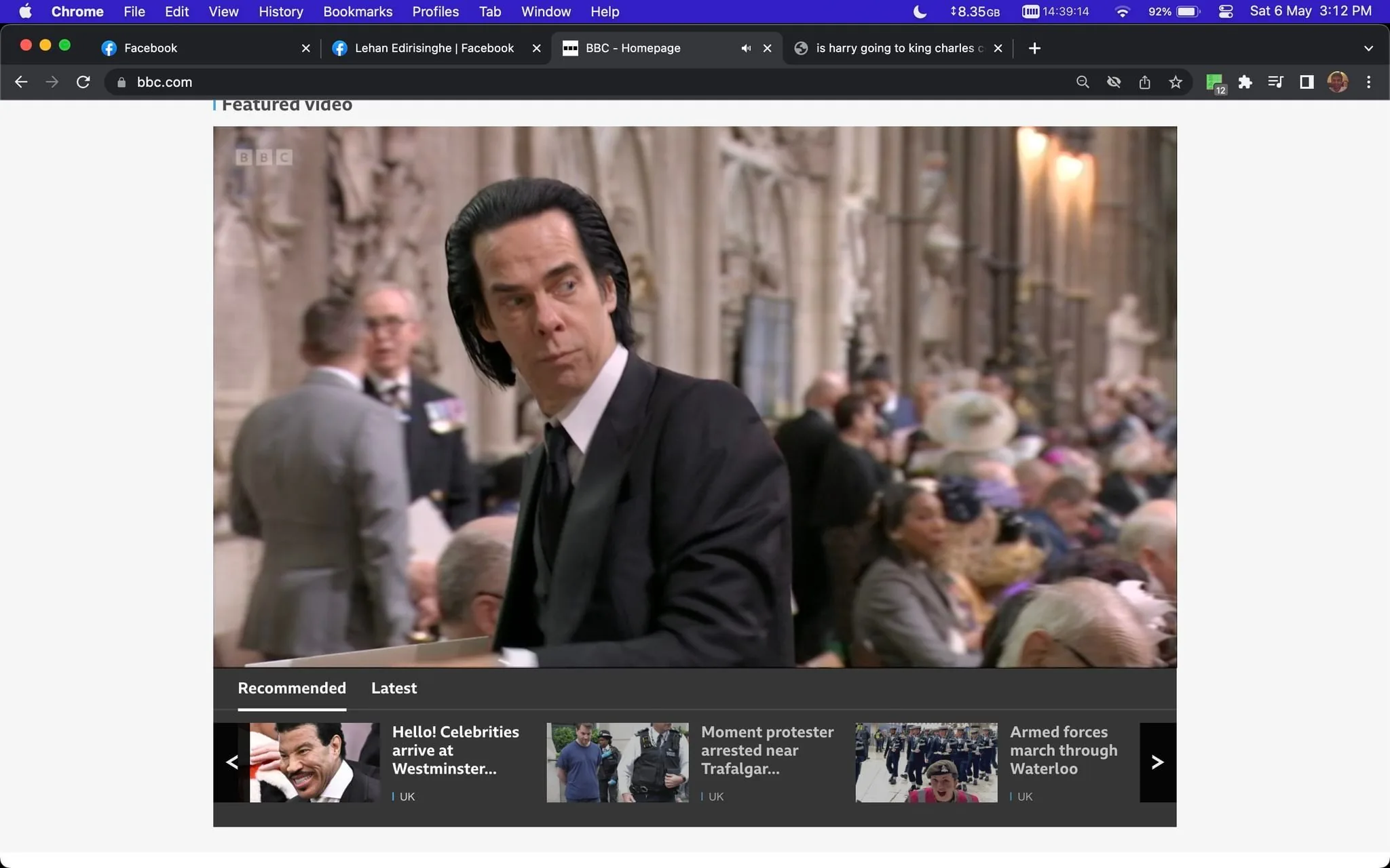 The image is a screenshot from BBC.com, showing a video frame with the title "Featured Video." The scene features Nick Cave, who is identified from the hint provided. He is dressed in a formal black suit and tie, situated in an indoor setting that appears to be a large, ornate hall, likely during a formal event or gathering. The background is filled with other attendees, some seated and some standing, indicative of a formal occasion.
Nick Cave is looking to his left with a neutral or contemplative expression. The lighting is bright, typical of an indoor event environment, and the decor of the hall includes detailed stonework and large windows. The foreground shows a blurred view of people, possibly indicating movement and a lively atmosphere.
The website interface includes a row of recommended and latest news items at the bottom, with headlines such as "Hello! Celebrities arrive at Westminster..." and "Moment protester arrested near Trafalgar..." suggesting coverage of significant events. The BBC's branding is visible on the top left of the video frame, reinforcing the source of the content.