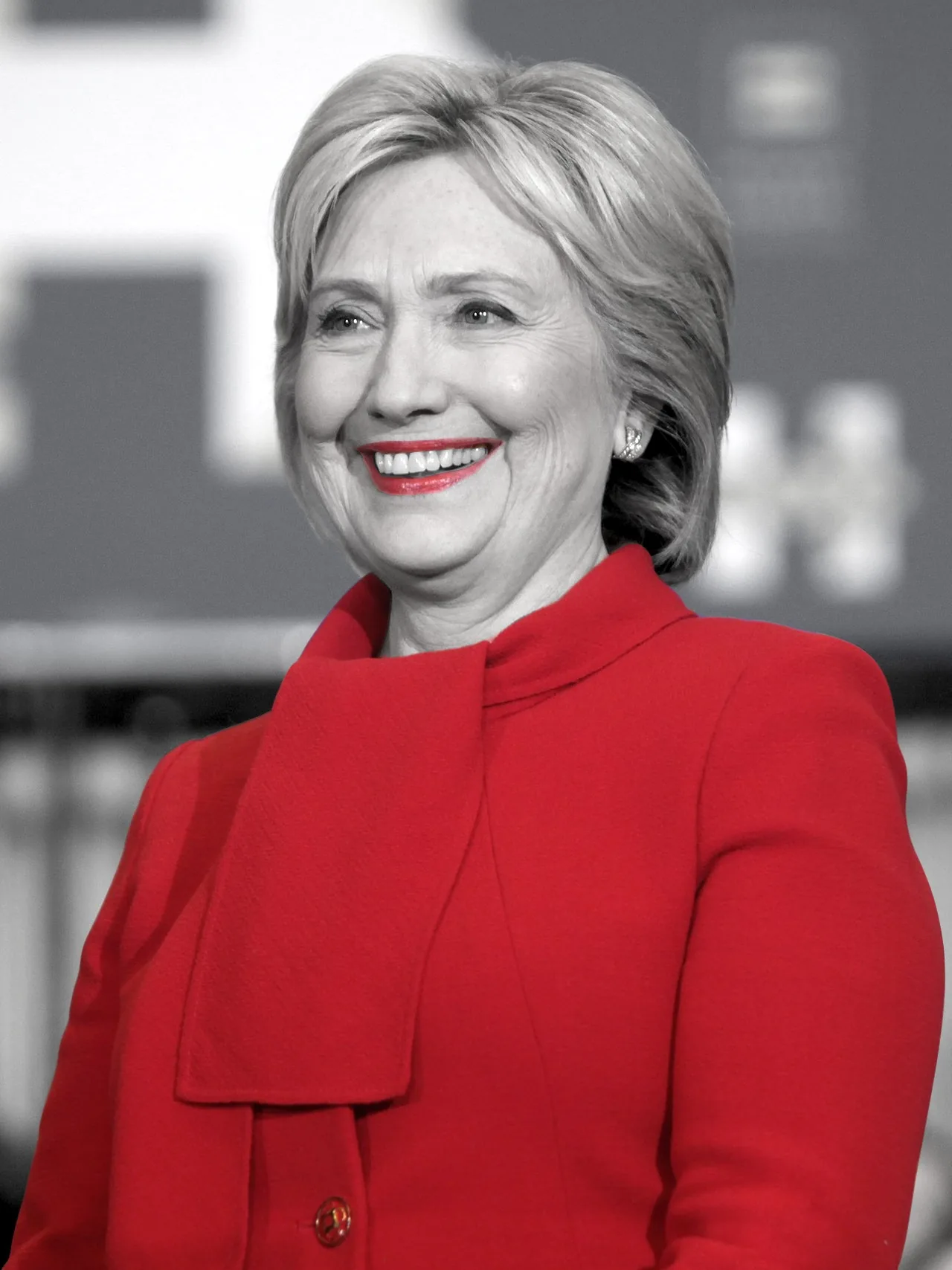 Hillary is depicted in this image with a striking color contrast. The photograph is primarily in grayscale, highlighting her in a vibrant red outfit, which creates a dramatic visual effect. Her attire, a formal red dress, captures attention with its boldness and elegance, standing out against the subdued background. Hillary is smiling warmly, suggesting a moment of success or celebration, indicating confidence and poise. Her hair is neatly styled, adding to her composed appearance. The blurred backdrop implies a public event or gathering, possibly a political or formal engagement. The lighting is soft yet focused, emphasizing her expression and the distinctive color of her outfit. This artistic choice of color accentuation symbolizes strength and triumph, aligning with the playful hint about "the blood of all the men she has defeated."