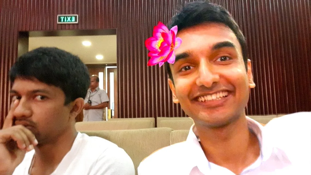 In this image, two individuals are seated indoors, likely at an event or gathering. On the right is Lehan Edirisinghe, who is smiling broadly and appears to be enjoying the moment. A digital flower filter adds a playful touch, positioned over his head with bright pink and red hues. On the left, Udhantha Sumathipala seems more reserved, with one hand touching his face, and a thoughtful expression. Both are seated in what appears to be an auditorium or conference room, characterized by wooden paneling and beige seating. The lighting is warm and creates a casual yet engaging atmosphere. A person is visible in the background, standing near an exit door marked with the word “EXIT” in bold letters. The image captures a moment of contrasting moods, with Lehan exuding joy and Udhantha appearing contemplative.