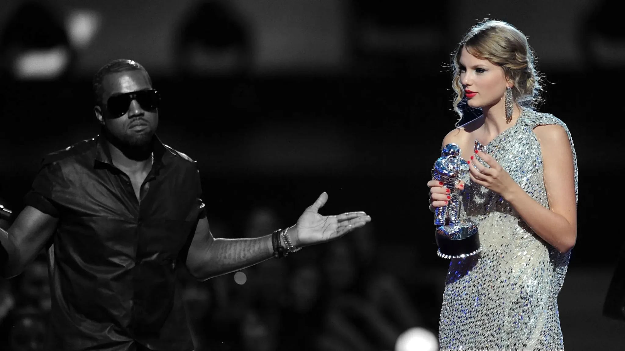 The image captures a notable moment during an award ceremony featuring Kanye and Taylor Swift. Kanye, dressed in a dark shirt and sunglasses, stands with an expressive gesture, arms outstretched. His demeanor appears assertive, possibly indicating he is speaking or interrupting. Taylor Swift, in a sparkling, silver gown, holds a trophy resembling an astronaut. Her expression is composed, though her posture suggests a moment of surprise or discomfort. The lighting focuses primarily on the two individuals, highlighting the drama of the scene. The background is out of focus, emphasizing the interaction between them at the forefront. This image is well-known for capturing a controversial moment during an award presentation.