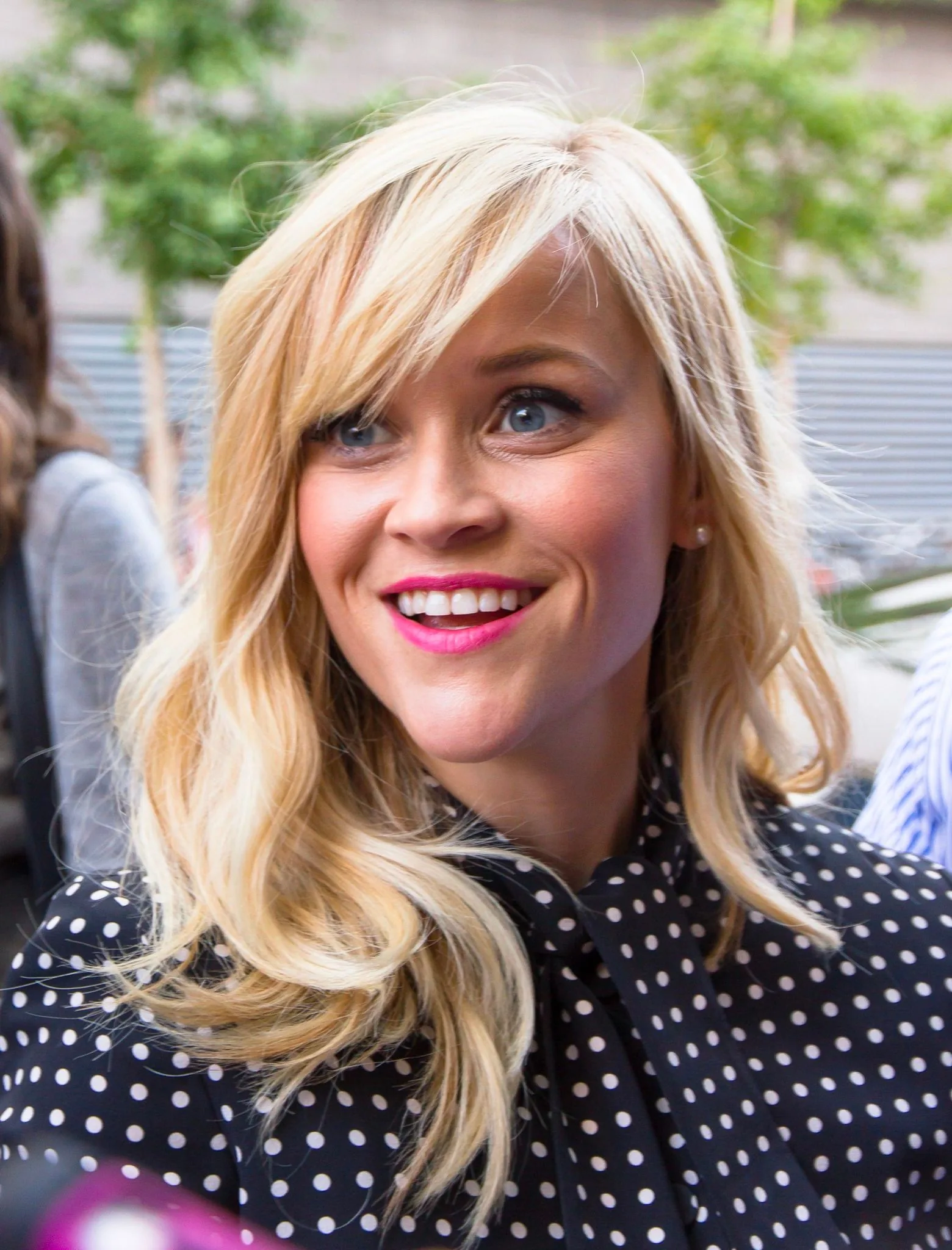 Reese Witherspoon is captured in this image, standing outdoors during the daytime. She is smiling brightly, which adds a lively and engaging atmosphere to the photograph. Her hair is styled in loose, wavy blonde locks that frame her face. She is wearing a black blouse adorned with white polka dots, providing a classic yet playful look. The background is slightly out of focus but features green foliage and a building, suggesting an event or public appearance. The lighting is natural and illuminates her features well, enhancing the vibrancy of her expression. The composition focuses primarily on Reese Witherspoon, with other figures blurred in the background, keeping the viewer's attention squarely on her. The overall mood of the image is cheerful and upbeat.