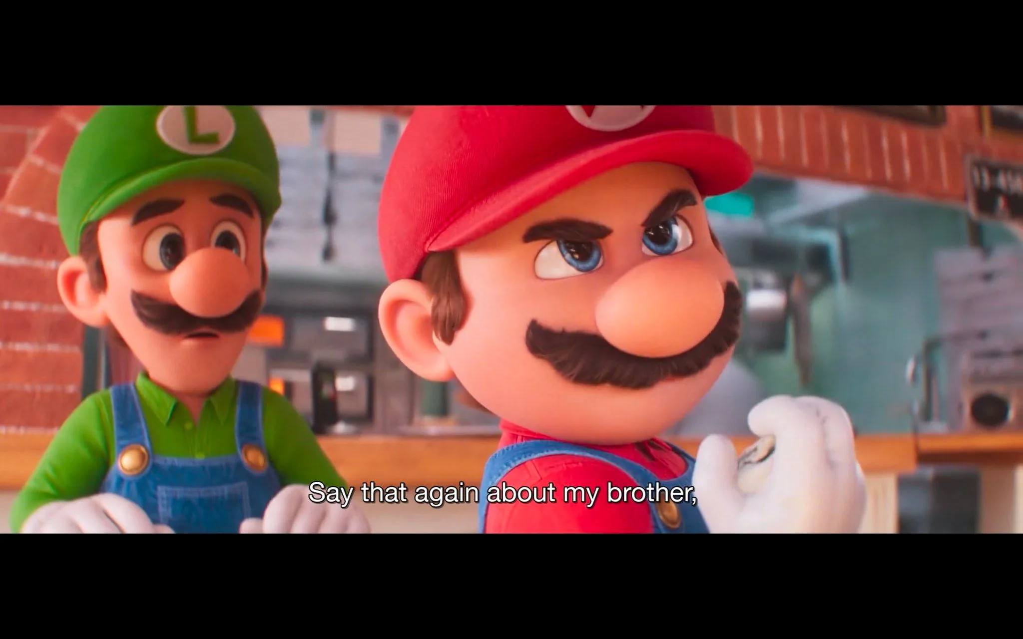 The image is a frame from an animated film featuring the well-known characters Mario and Luigi from the "Super Mario" franchise. In this scene, Mario is in the foreground wearing his iconic red cap and blue overalls, with a stern expression as he clenches a fist, suggesting he's either angry or ready to defend someone. Luigi, standing slightly behind, appears concerned or anxious, dressed in his usual green cap and blue overalls. The setting seems to be indoors, possibly a casual environment like a kitchen or workshop, given the visible brick walls and stainless steel appliances in the background. The lighting is bright, enhancing the vivid colors of the characters' outfits. The style is typical of modern 3D animation, with detailed textures and expressive faces. The subtitle reads, “Say that again about my brother,” indicating a protective or confrontational moment involving Luigi. This snapshot captures the dynamic and loyal relationship between the two brothers.
