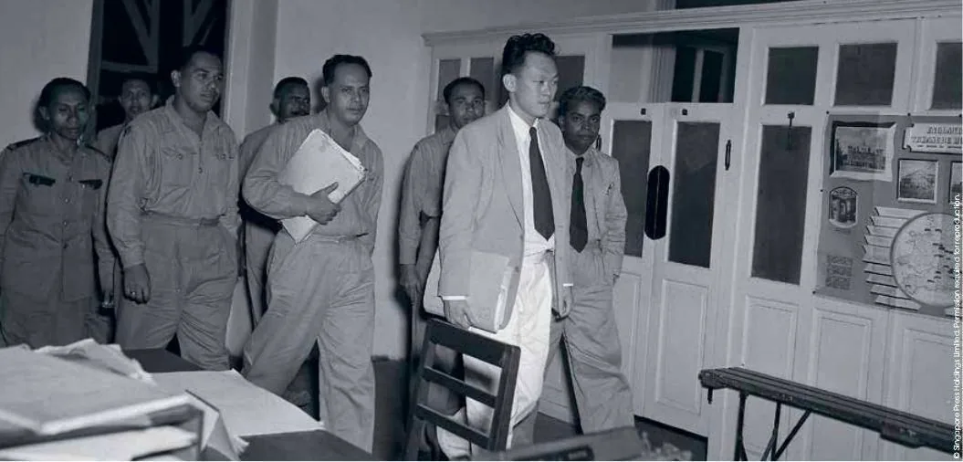 The image depicts a formal setting, likely a meeting or significant gathering. In the center of the image, a man identified as Lee Kuan Yew leads a group of individuals. He is dressed in a light-colored suit with a dark tie, exuding a serious and focused demeanor. The other men in the scene are wearing uniforms, suggesting a military or official context. They follow closely behind, some carrying documents, which indicates a meeting or briefing might be occurring.
The room is furnished with desks and chairs, and there are various papers scattered on a desk in the foreground, enhancing the professional atmosphere. The walls are adorned with notices and posters, and a partially visible Union Jack flag suggests a colonial or diplomatic environment. The lighting is bright and even, typical of indoor office settings.
The text in the image mentions "Singapore Press Holdings Limited. Reprinted with permission," indicating the image is sourced from a reputable archive or publication. This historical photograph captures an important moment, likely involving discussions or decisions of political significance.