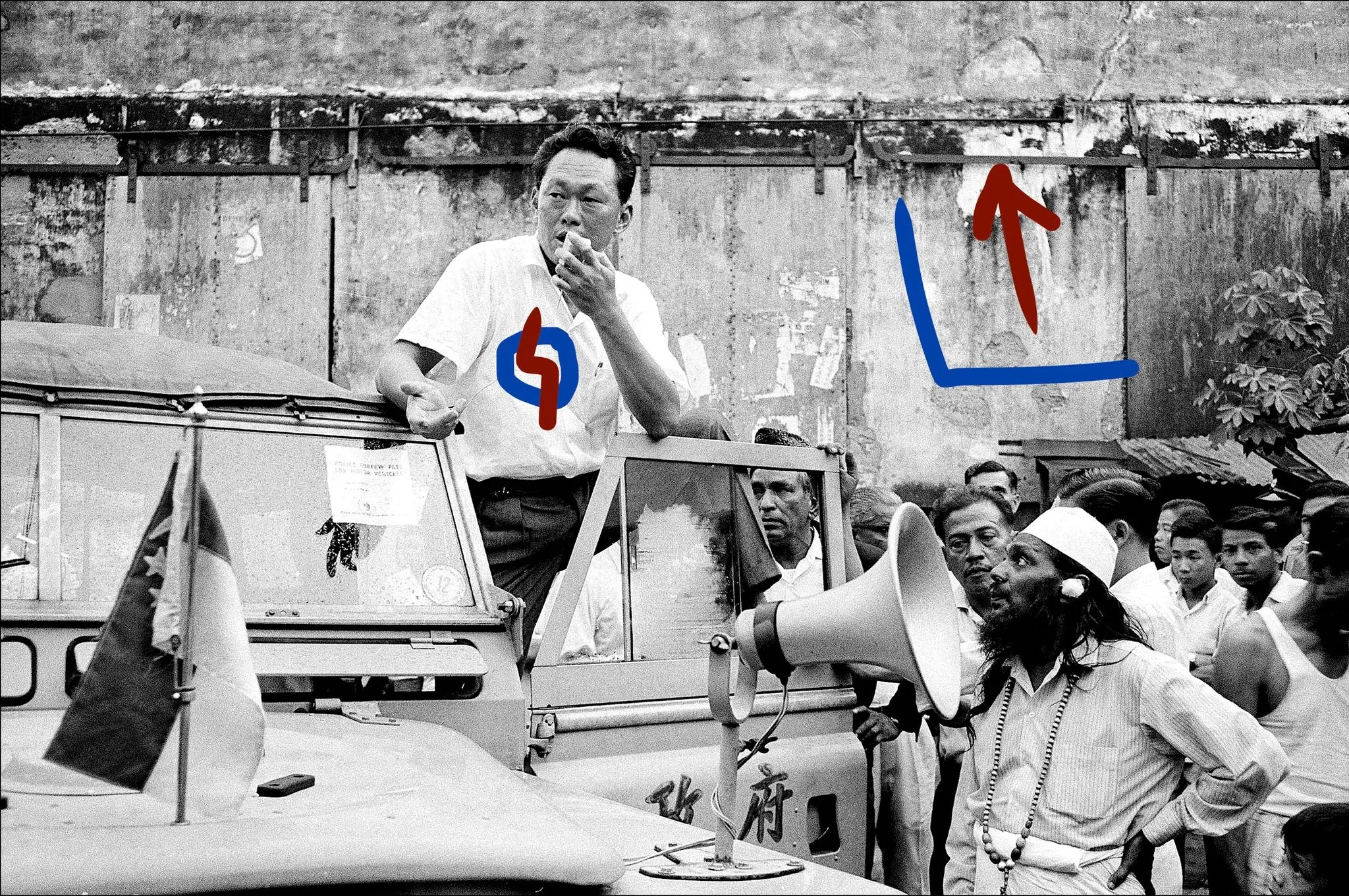Lee Kuan Yew is prominently featured in this historical black-and-white photograph. He is standing on a vehicle, possibly a jeep, with a microphone in front of him, suggesting he's delivering a speech or addressing a crowd. The scene seems to capture a political event or campaign rally, evidenced by the presence of flags and a large megaphone. Lee Kuan Yew wears a short-sleeved shirt, and his posture is dynamic, indicating engagement with the audience. Surrounding him are several individuals, one of whom appears to be a man in traditional attire with a beard and a cap. The backdrop is a worn-out wall, adding a rustic element to the setting. The scene is bustling with activity, as people in the crowd appear attentive and focused on the speaker. Text visible in the image includes "POLICE TOW AWAY IN FORCE" on the vehicle and some Chinese characters on the side panel. The overall mood of the image reflects political fervor and public engagement.