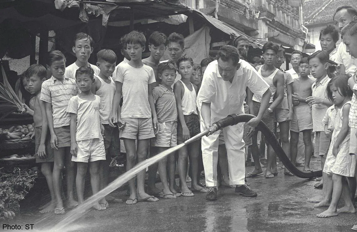 In this historical black-and-white photograph, a group of children and adults are gathered in an outdoor setting, likely a market or street in Singapore. The focal point of the image is Lee Kuan Yew, who is actively engaged in spraying water from a hose onto the street. He is wearing a white shirt and pants, and is bent over slightly, focused on the task at hand. The children surrounding him are dressed in casual attire typical of the era, with shorts and simple tops. Their expressions range from curiosity to fascination as they watch the water flow from the hose. The background shows modest structures with a market-like atmosphere, including visible produce. The scene captures a moment of communal activity, reflecting a sense of community engagement and leadership. The photograph is credited to "ST."