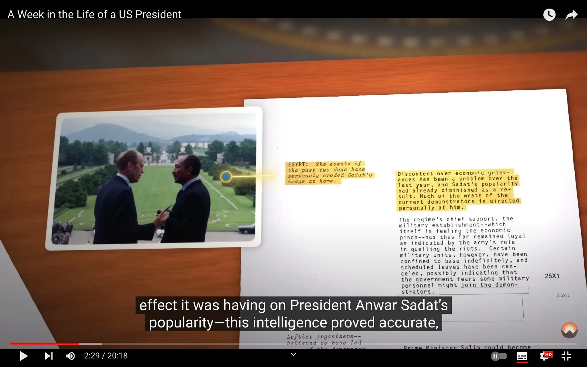 The video frame, titled "A Week in the Life of a US President," features a photograph juxtaposed with a document on a wooden table. The photograph shows two men standing outdoors, seemingly engaged in conversation, with a scenic, well-manicured lawn and distant hills in the background. The man on the left holds a pen, while the man on the right, identified in the context as President Anwar Sadat, appears to be smiling. Both are dressed in formal suits, suggesting a diplomatic or official meeting.
Beside the photograph, a document contains highlighted text discussing the political situation in Egypt and President Sadat's waning popularity due to economic grievances. Highlighted sections of the text indicate that recent events have negatively impacted Sadat’s image at home.
The atmosphere conveyed is diplomatic and tense, drawing attention to the delicate nature of international relations. The lighting is bright, reflecting a clear day, and the overall composition suggests a historical or political analysis.