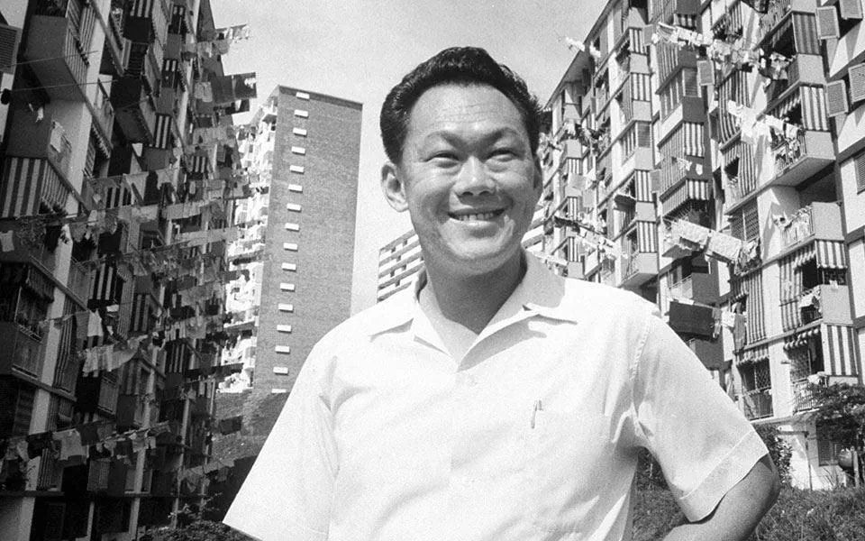 The image is a black and white photograph of Lee Kuan Yew, an influential figure in Singapore's history. He is seen standing outdoors, smiling broadly, suggesting a positive or confident demeanor. Lee is dressed in a simple, short-sleeved white shirt, which adds to the informal and approachable aura of the photo. The background features high-rise apartment buildings, indicative of urban development. Clothes are hanging from the balconies, a common sight in densely populated areas, hinting at the everyday life of residents. The composition captures a juxtaposition of human expression and urban architecture, highlighting both personal and societal growth. The lighting is natural, suggesting the photo was taken during the day. This image likely captures a moment of pride or achievement in urban development or public housing projects.